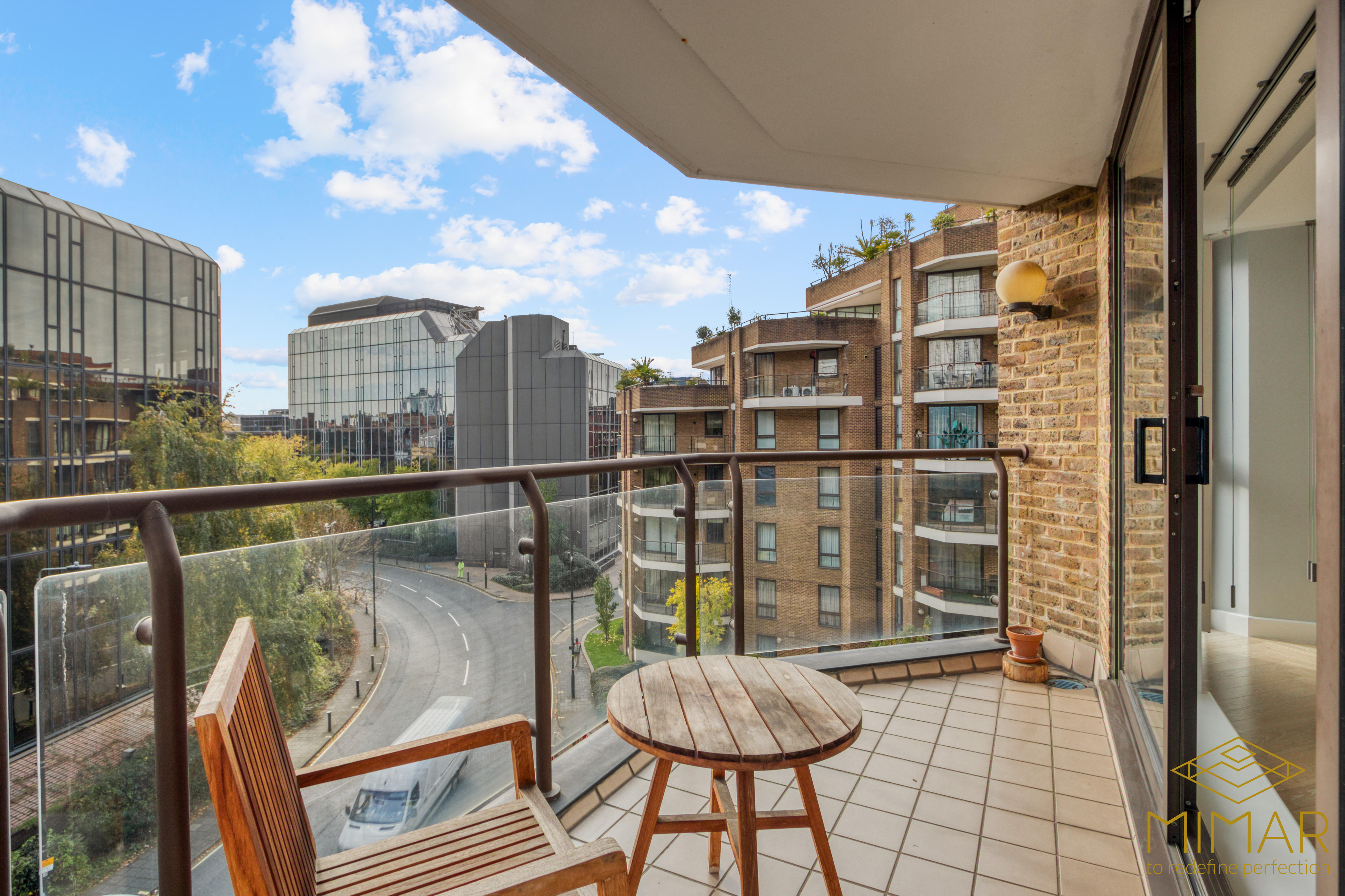 A modern apartment balcony with glass railing, wooden chair and table, overlooking urban buildings.