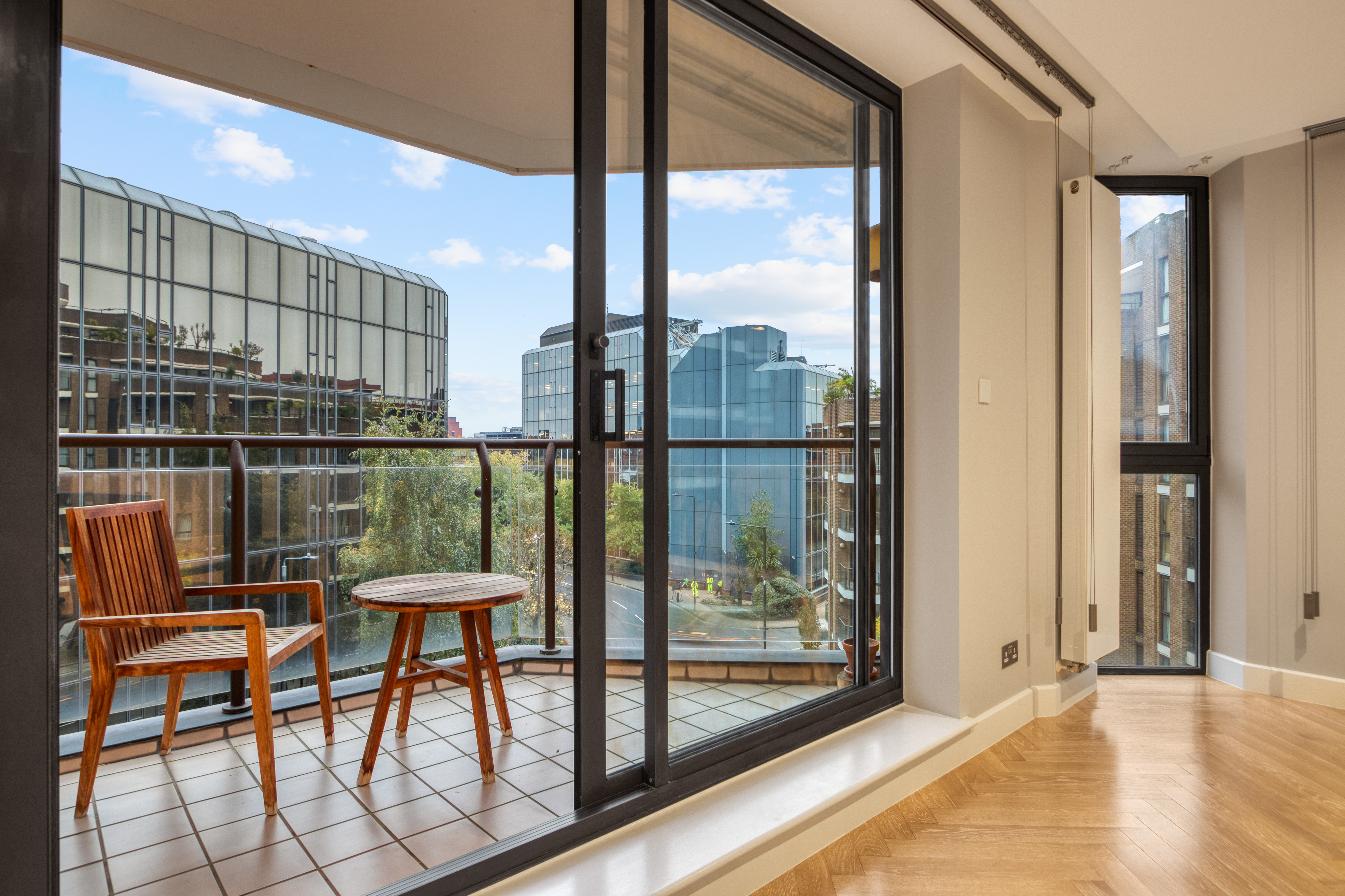 A modern loft conversion with a glass sliding door opening to a balcony, featuring wooden chairs and a table, overlooking urban buildings.