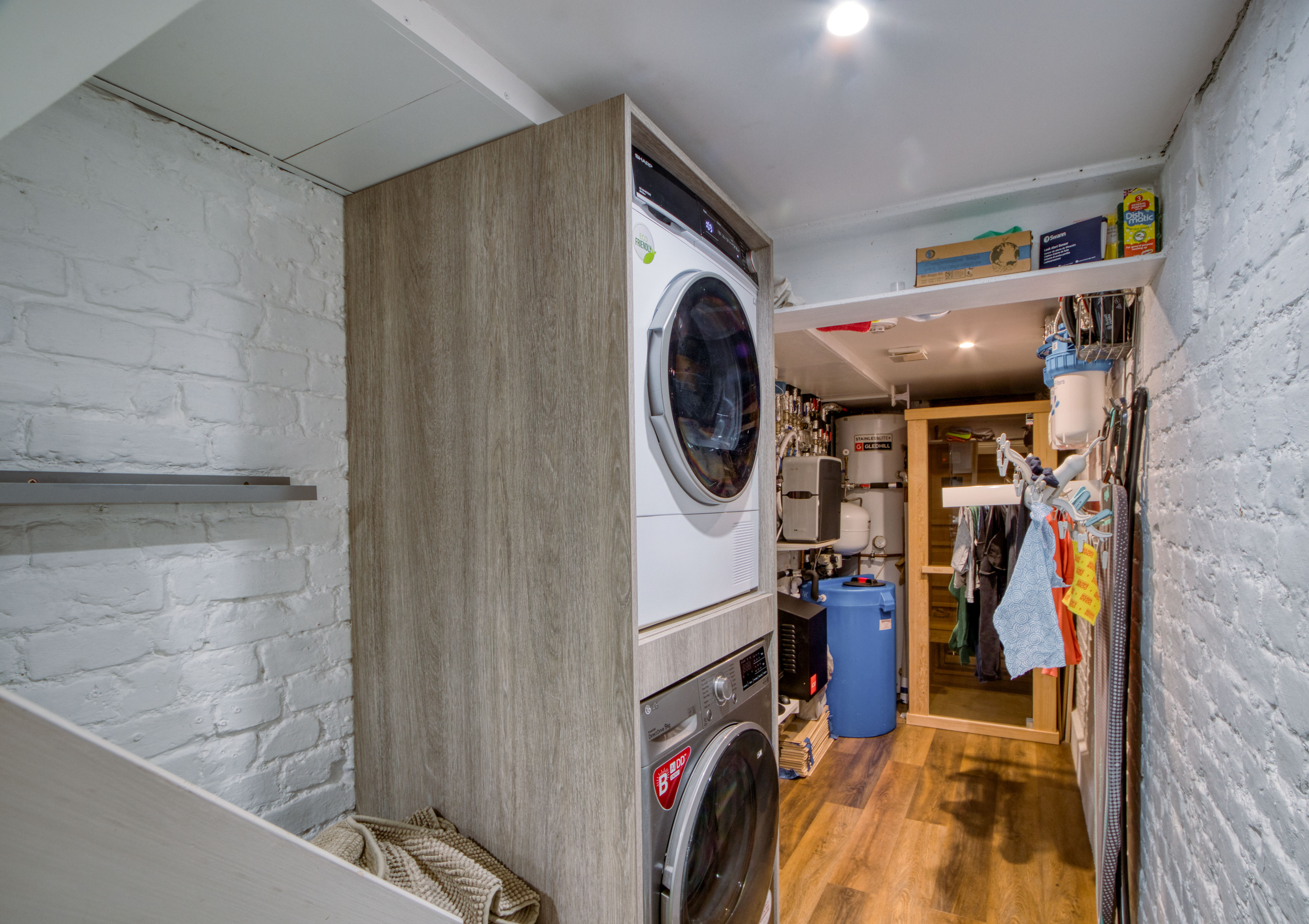 The image shows a well-organized basement laundry room with stacked washer and dryer units, wooden flooring, and an exposed brick wall. Various plumbing and utility components are visible.