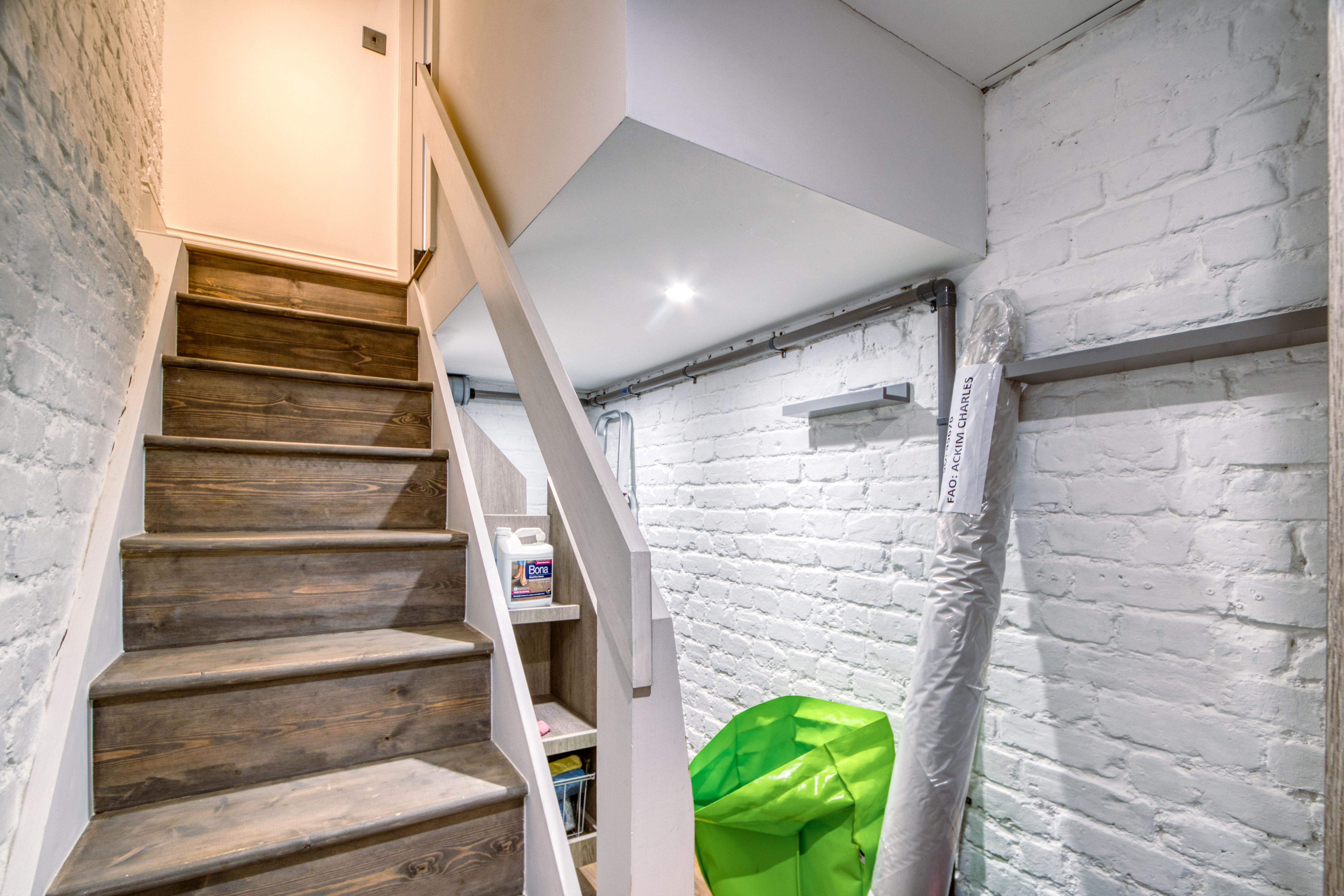 A newly renovated basement area featuring a wooden staircase leading upwards, with white painted brick walls and storage shelves to the side.
