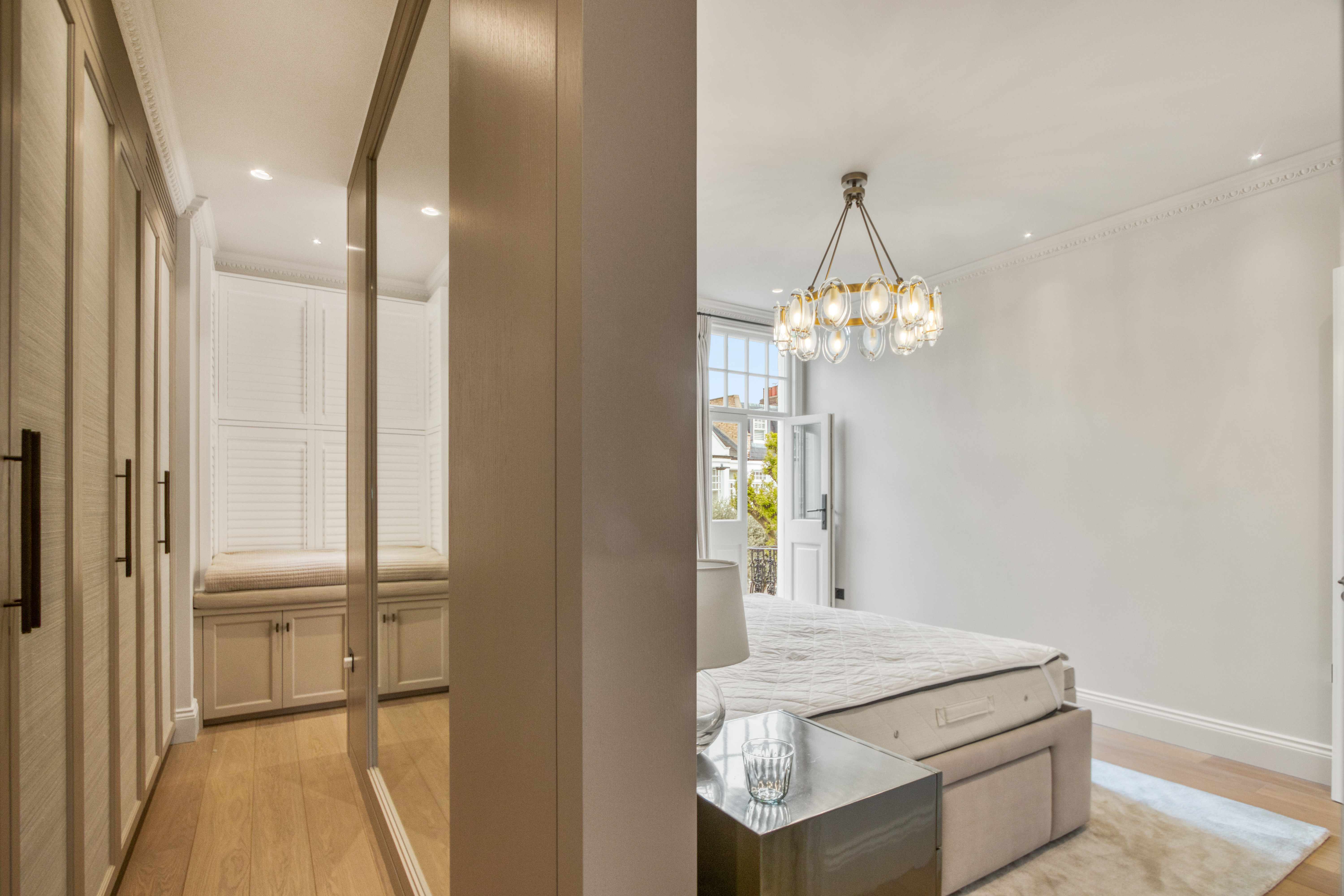 A modern refurbished bedroom with a visible walk-in closet on the left, featuring a chandelier, large windows, and an adjacent mirrored closet area.
