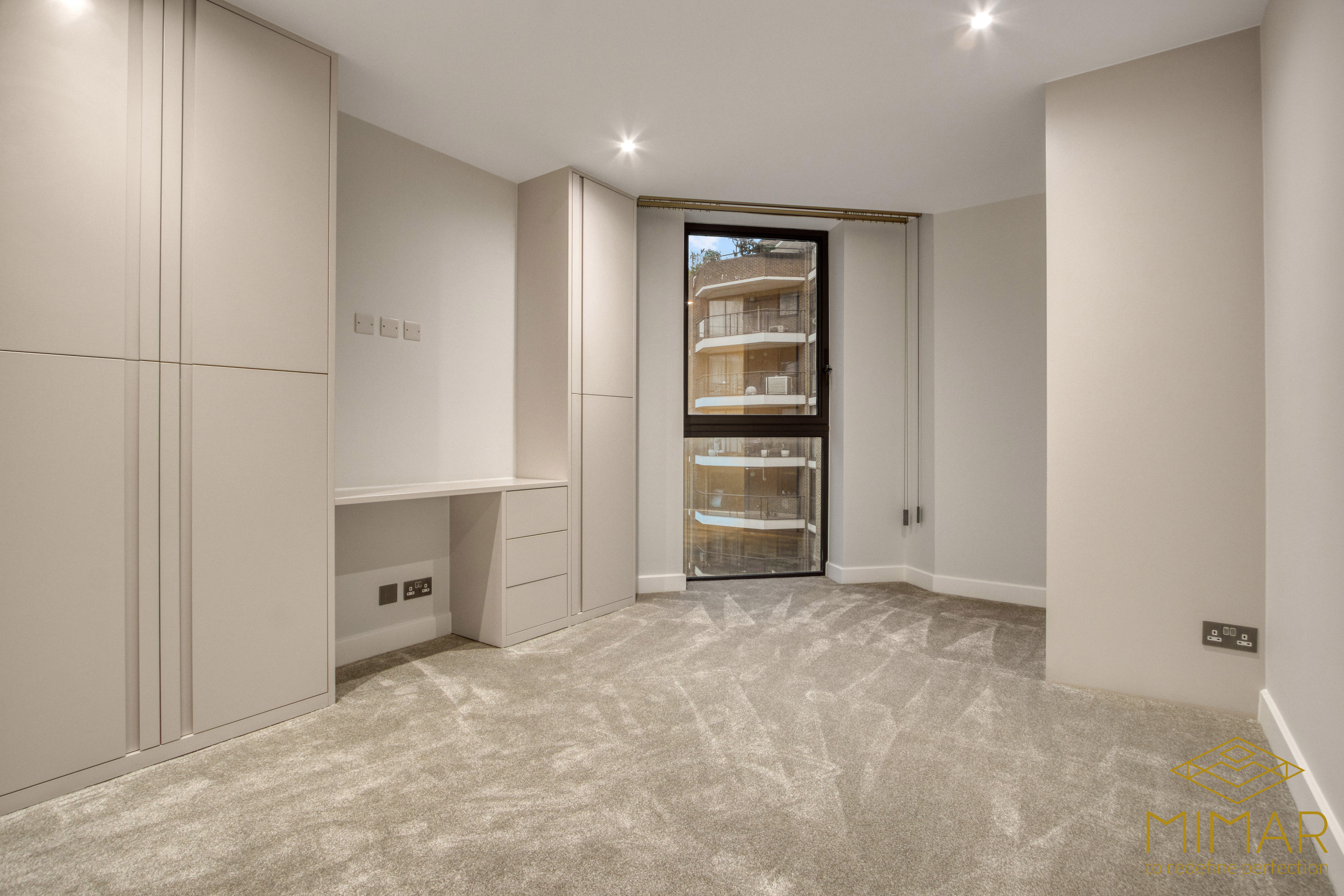 A modern bedroom interior showcasing custom built-in wardrobes and a small desk area with neutral coloring and a large window.