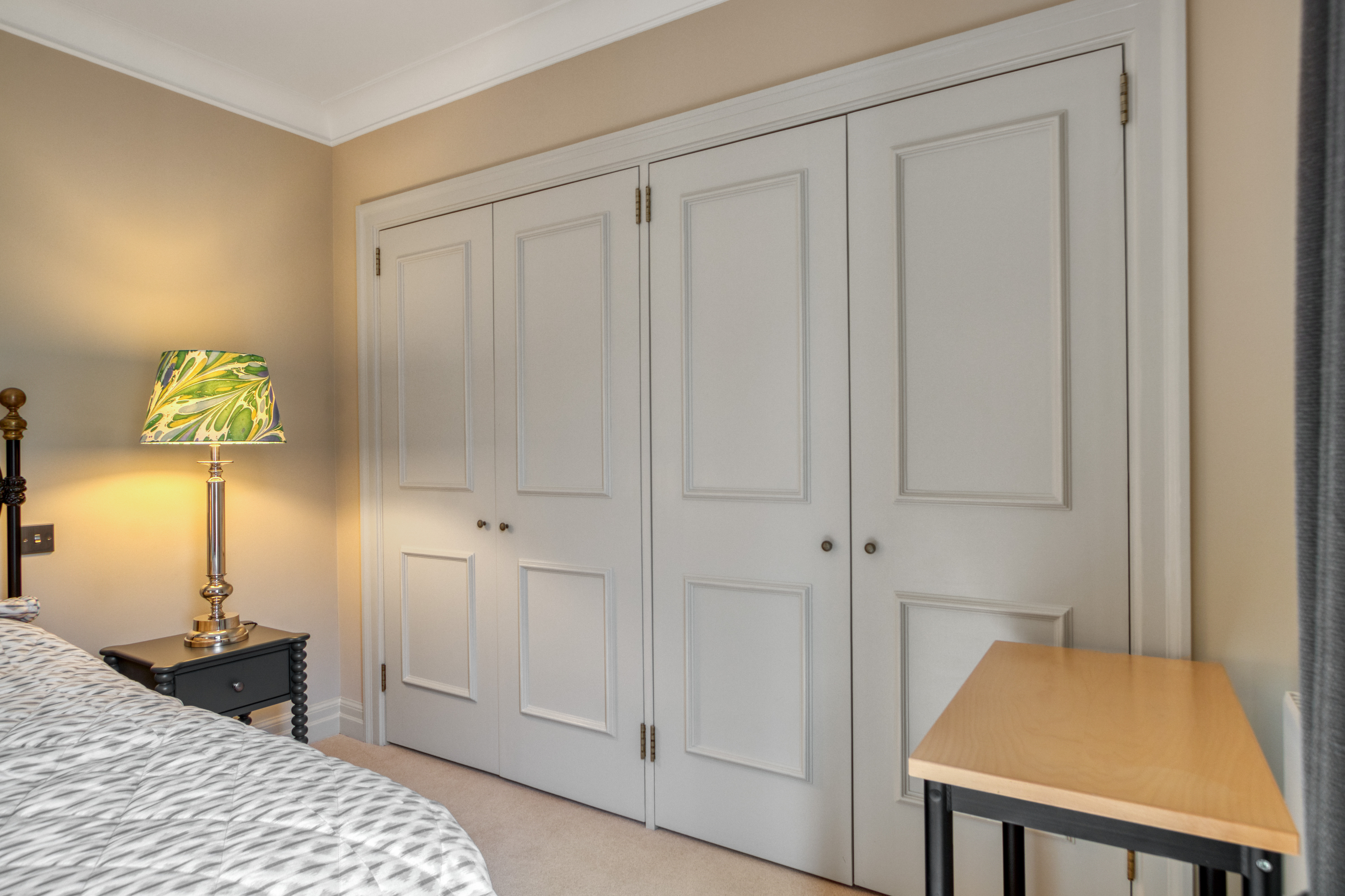 This image shows a bedroom featuring a custom-built wardrobe with double doors and a bedside lamp on a table.