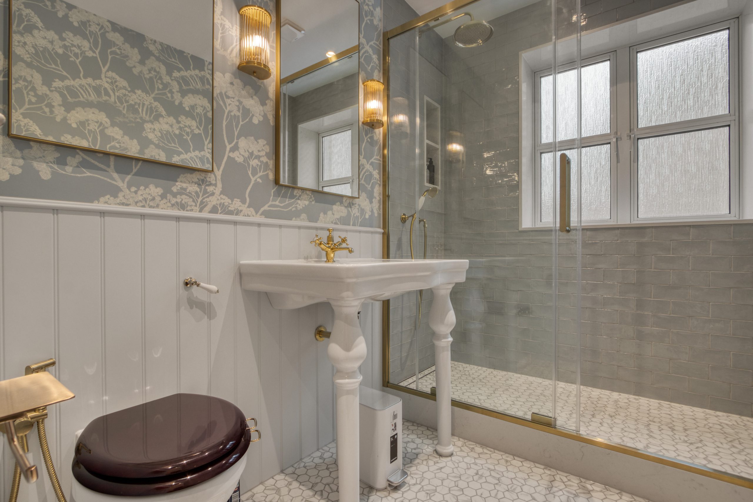 A luxurious bathroom featuring a white pedestal sink, gold fixtures, a glass-enclosed shower, patterned wallpaper, and tiled flooring.