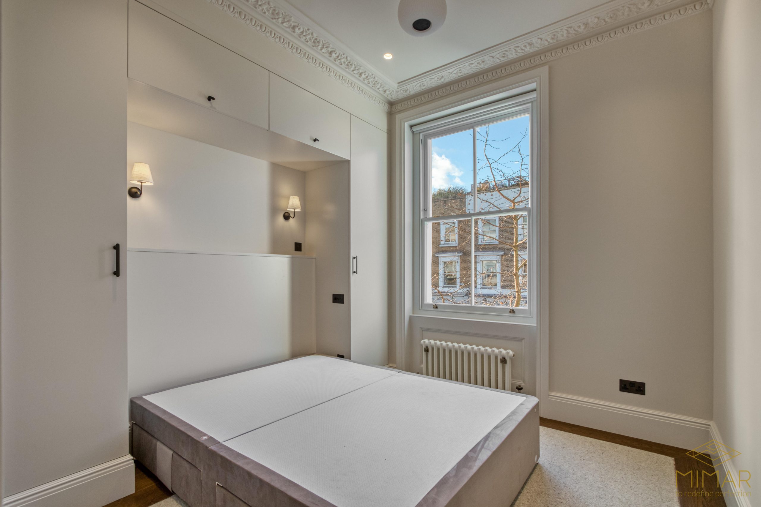 A remodeled bedroom featuring built-in wardrobes, ceiling moldings, a radiator, and a large window with a view of neighboring houses.