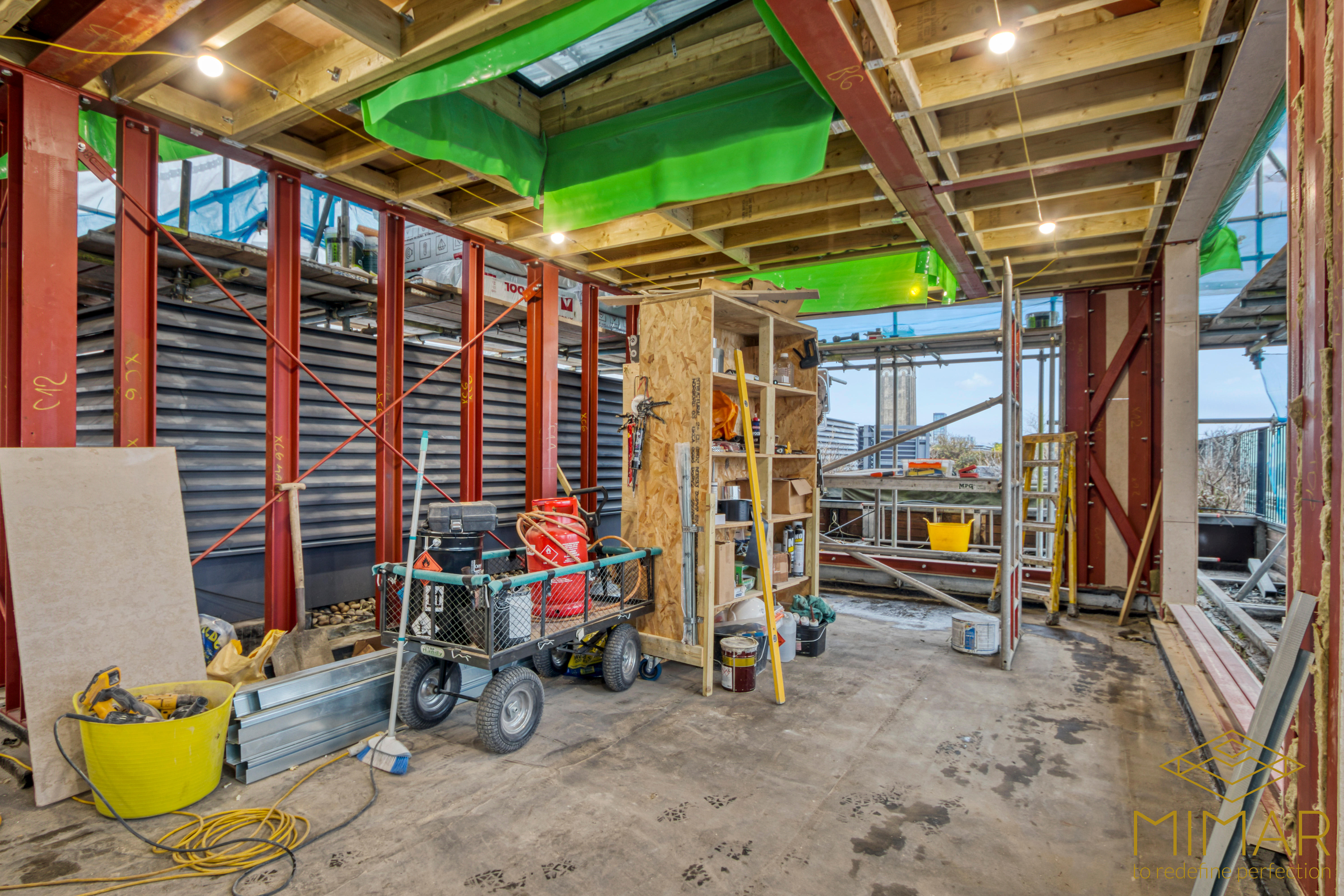 Interior view of a house extension under construction with exposed steel beams, wooden framework, and construction materials on site.