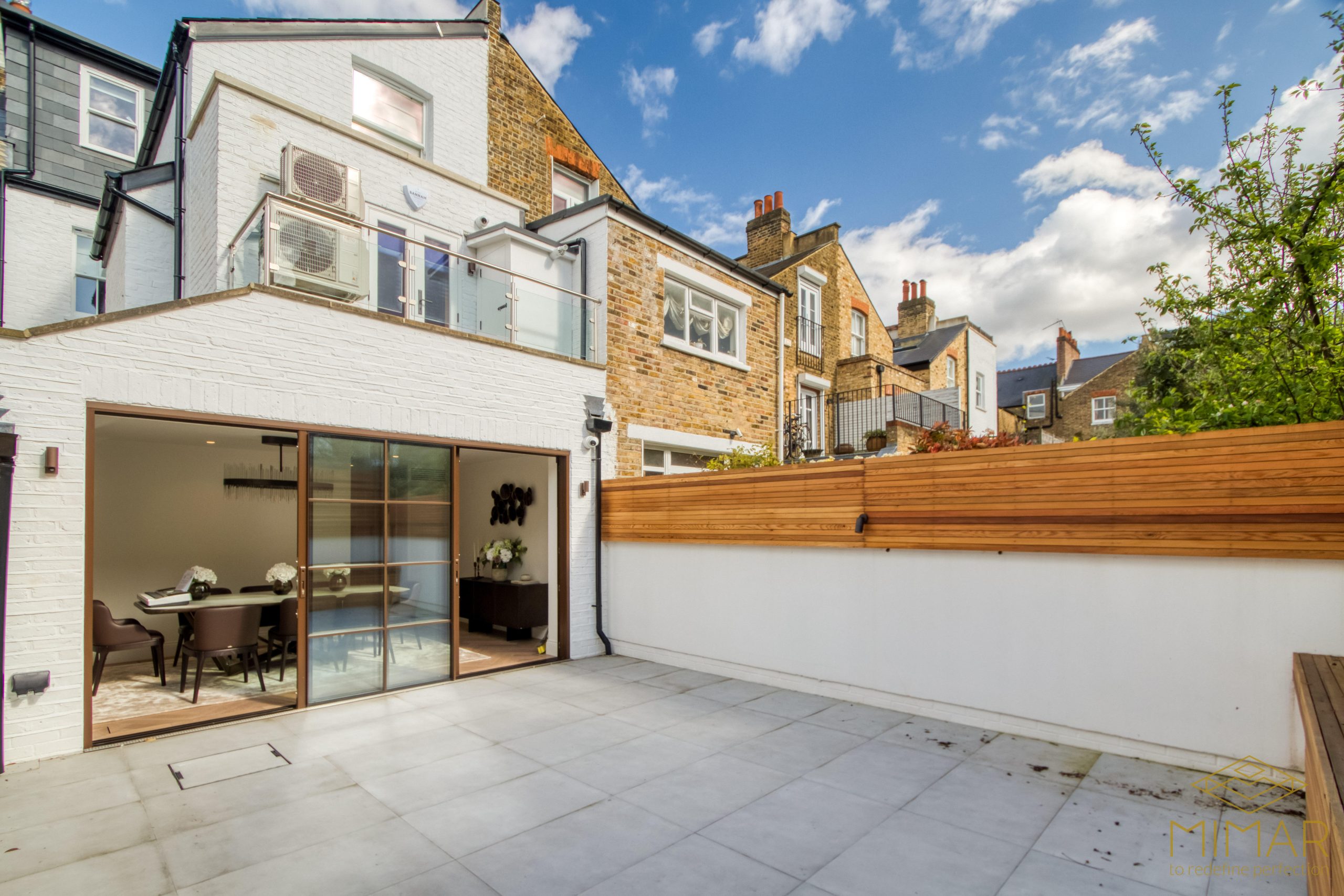 Modern house extension with a glass-door entrance to a dining room, featuring a spacious tiled patio area and wooden privacy fence.