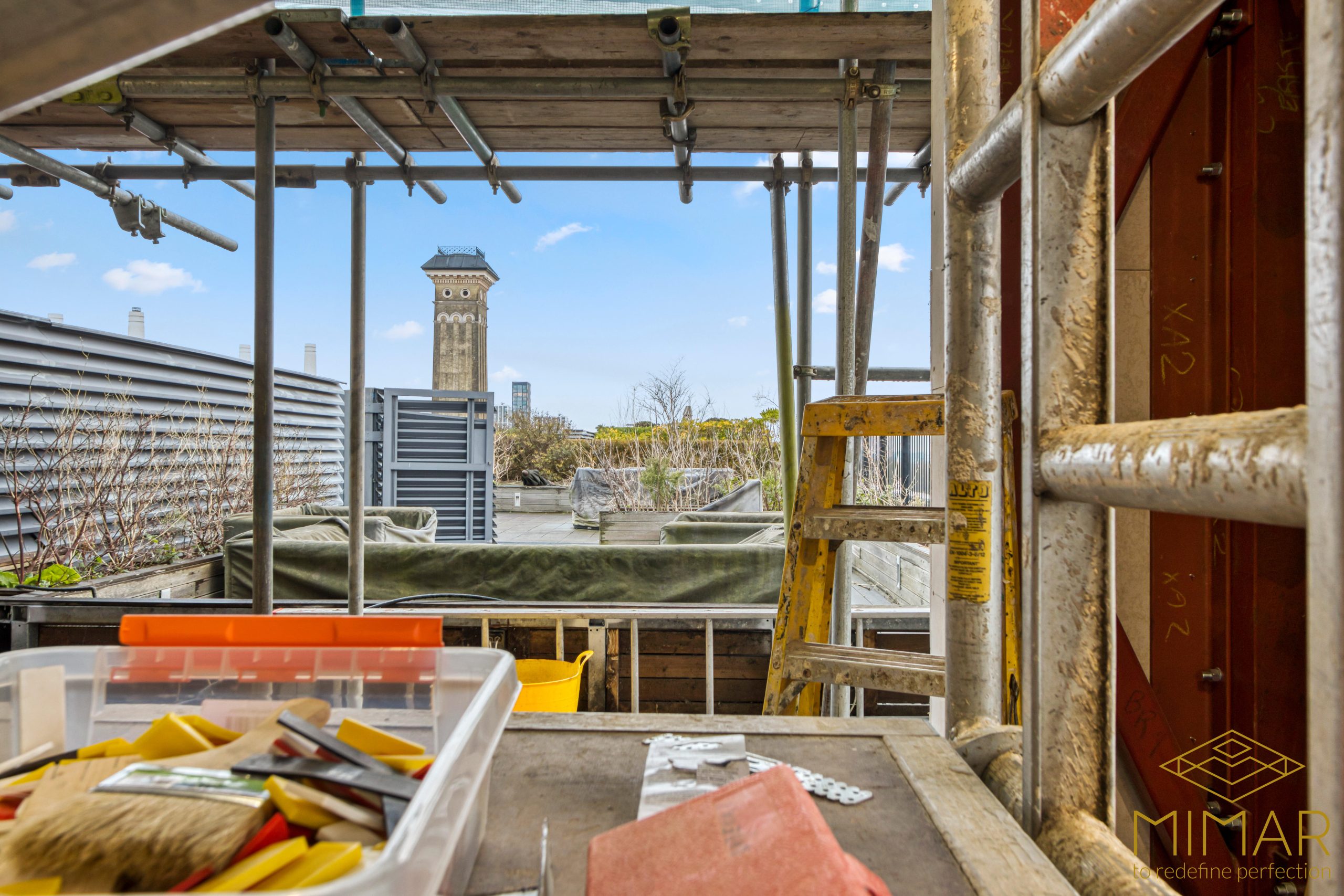 The image shows a construction site with scaffolding, a ladder, and various tools on a table, overlooking a cityscape with a tall building in the distance.