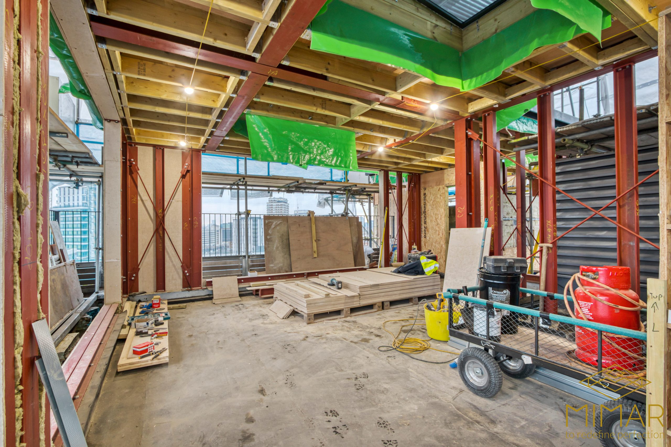 Interior construction site with exposed beams, metal structures, and construction materials inside a room with large windows.