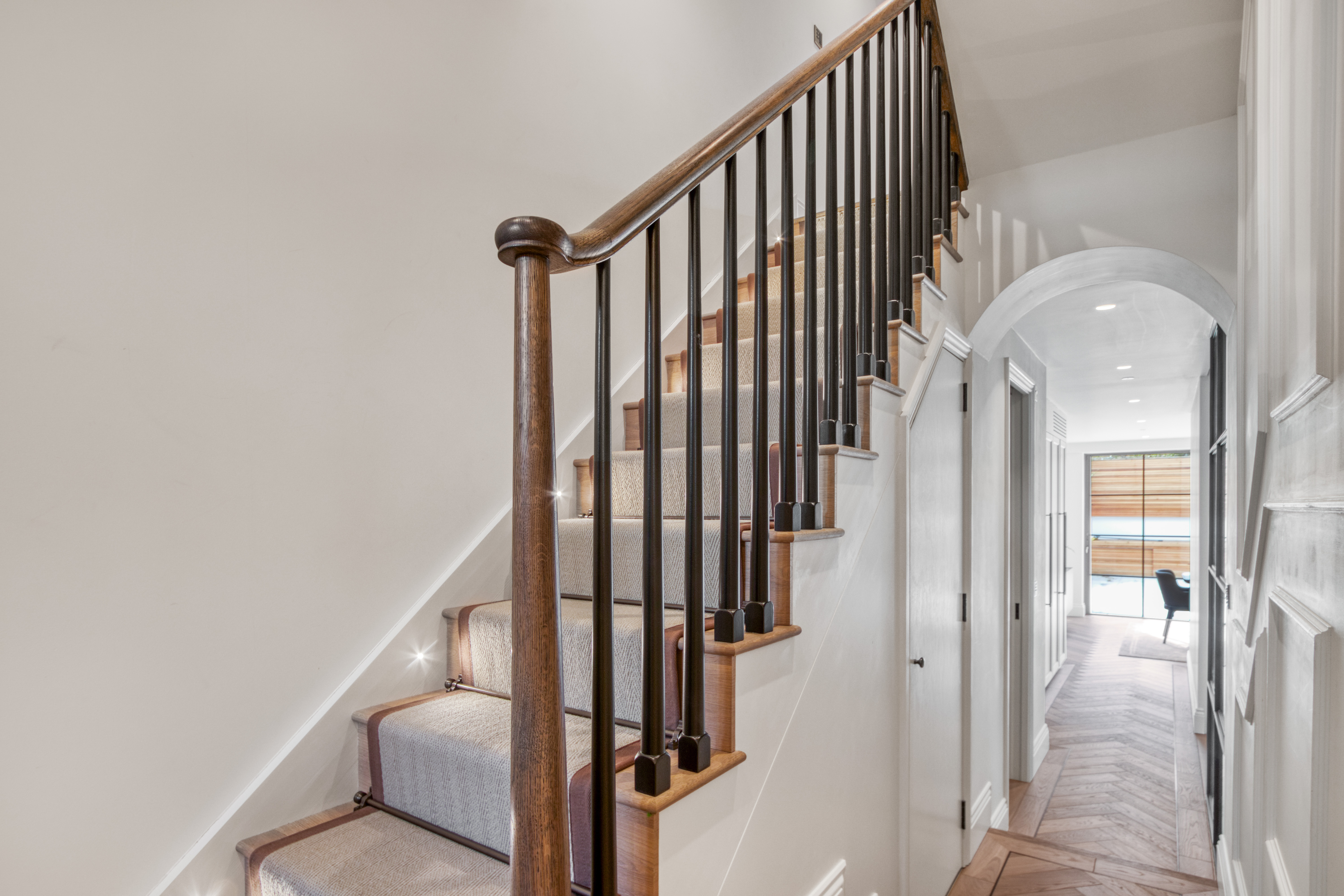A modern interior featuring a refurbished staircase with wooden handrails and carpeted steps, leading to an elongated hallway with natural lighting.