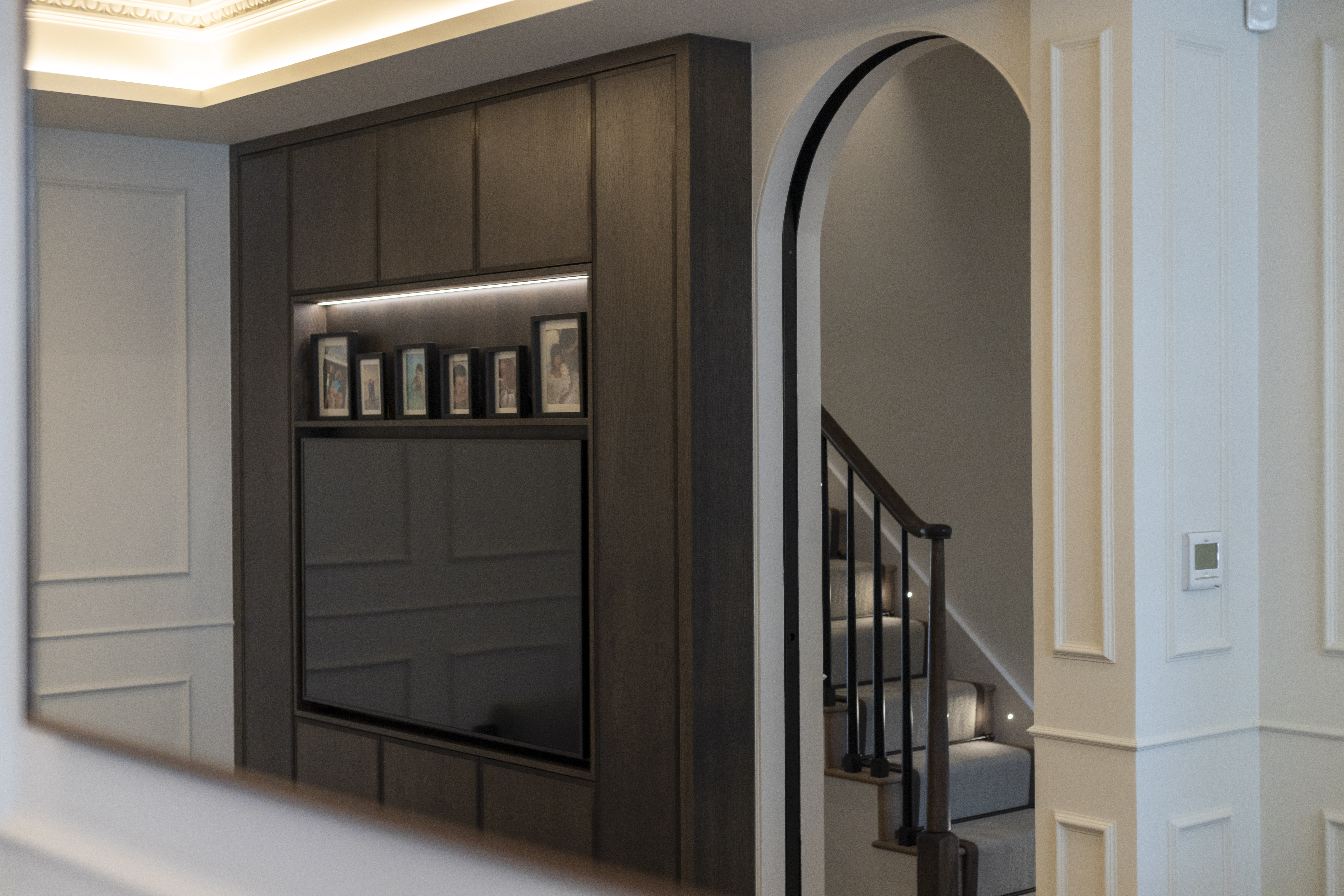 The image showcases a living area with a custom wooden accent wall featuring built-in lighting and photo frames, beside a staircase with modern railing.