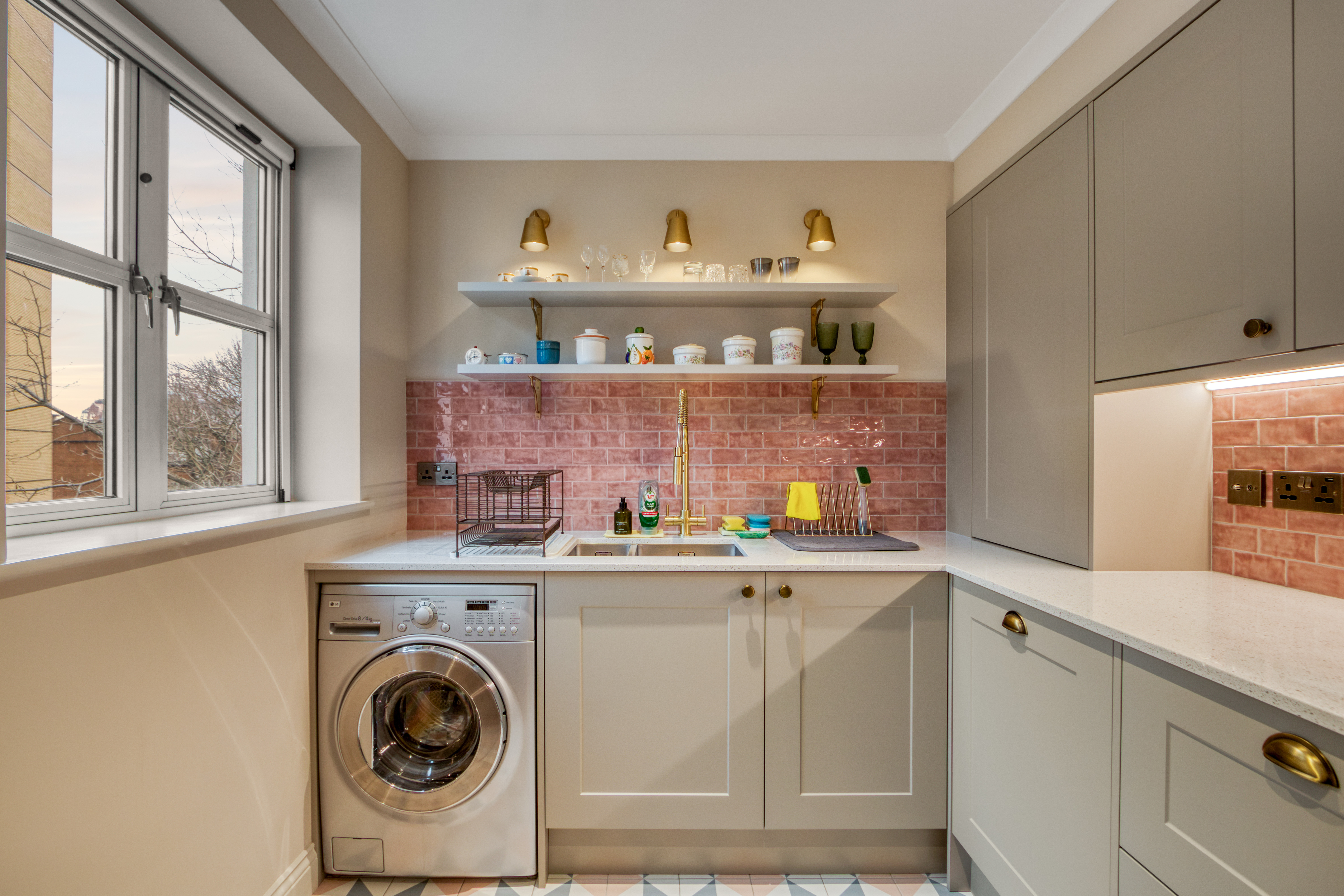 A modern kitchen renovation featuring sleek cabinetry, an integrated washing machine, open shelving, and stylish lighting with a pink tiled backsplash.