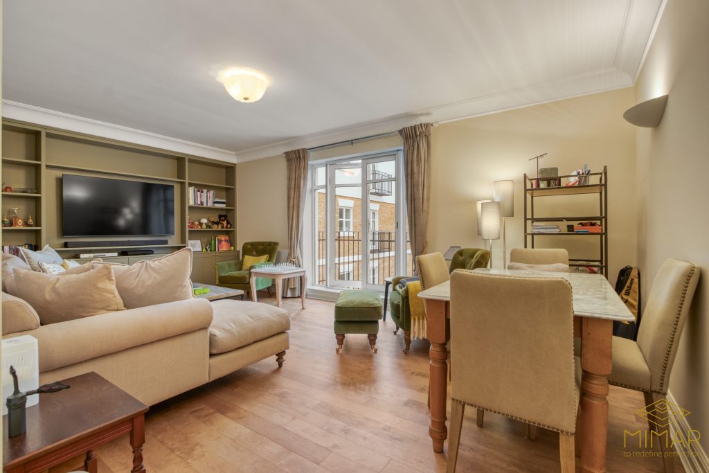 A modern living room with a large sofa, built-in shelving with a television, and a small dining table with chairs, featuring wooden flooring and a view out to a balcony.
