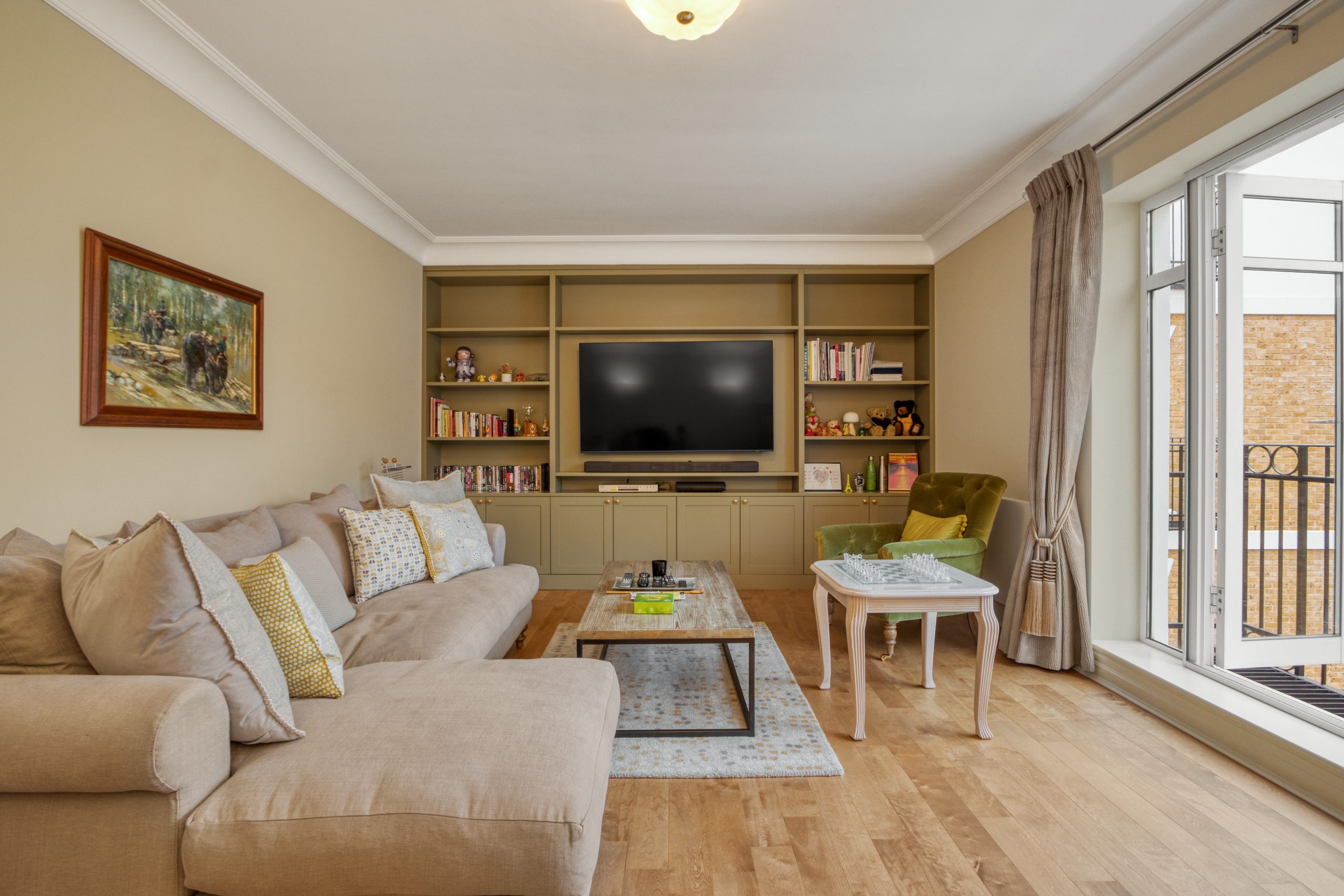 A remodeled living room with beige sectional sofa, custom shelving, and a mounted TV, featuring hardwood flooring and large windows.