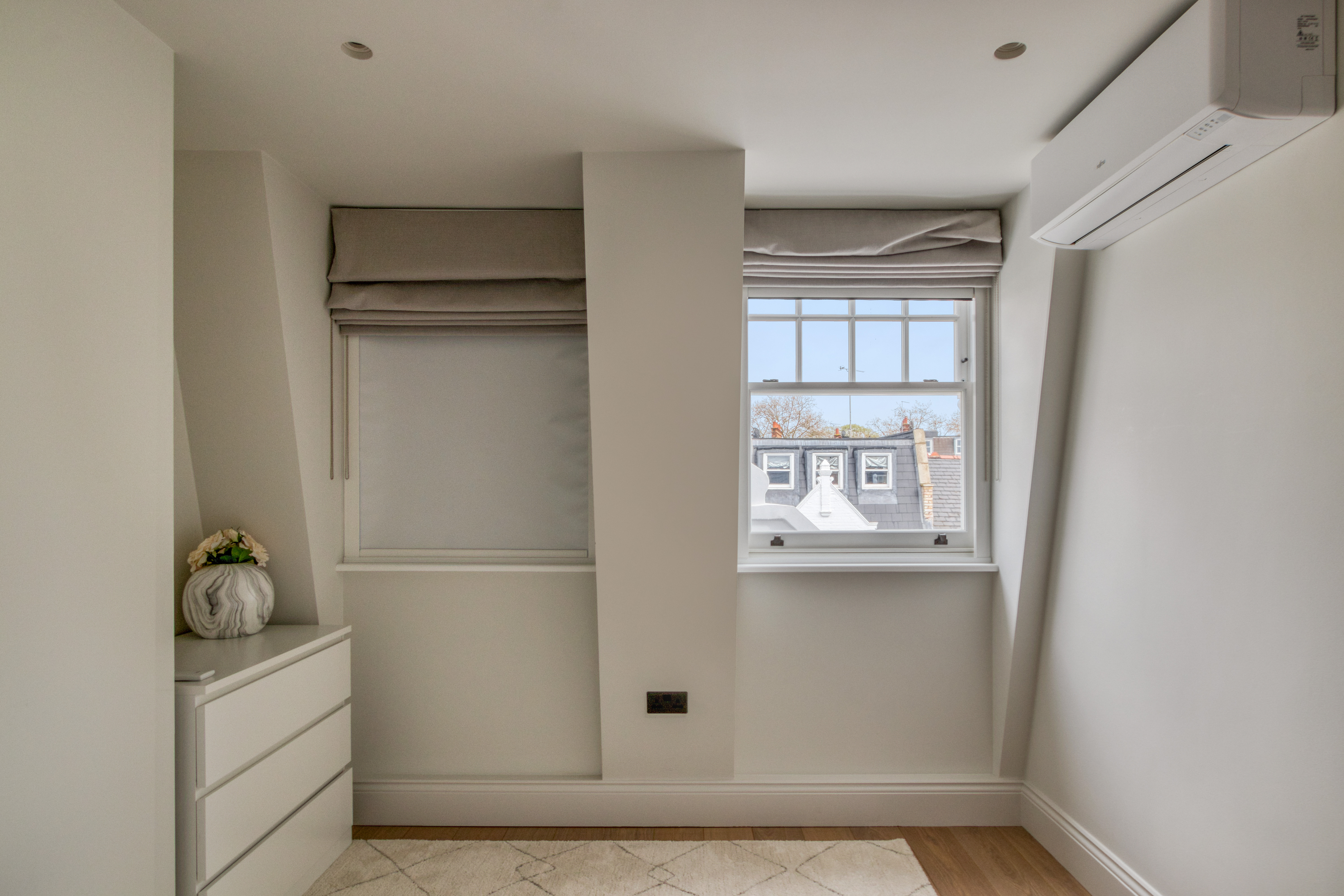 A loft bedroom with a dresser, two windows with blinds, and a white air conditioning unit mounted on the wall.