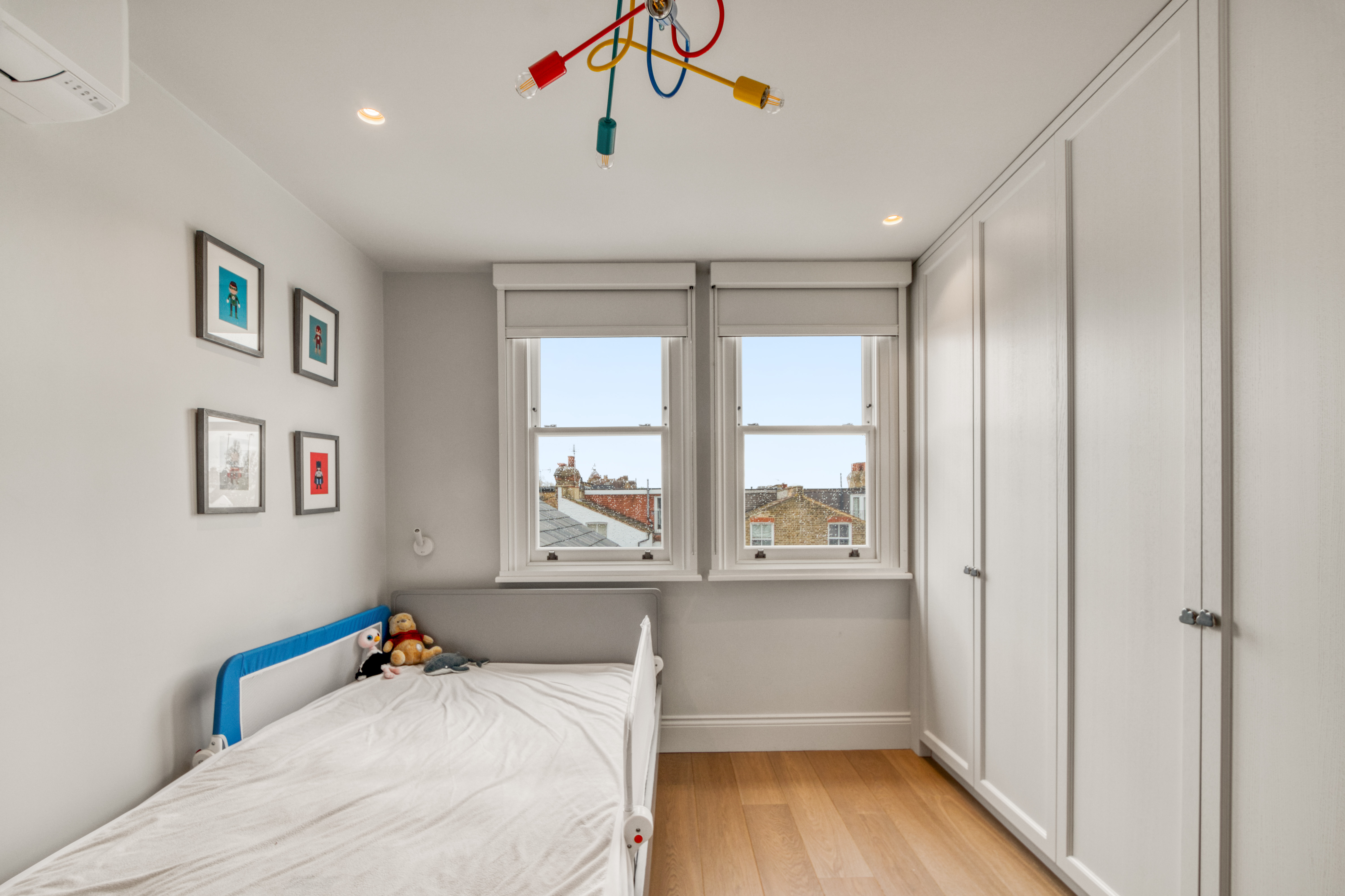 A modern loft bedroom featuring a single bed, built-in wardrobe, and colorful pendant light with wooden flooring and exterior views through windows.