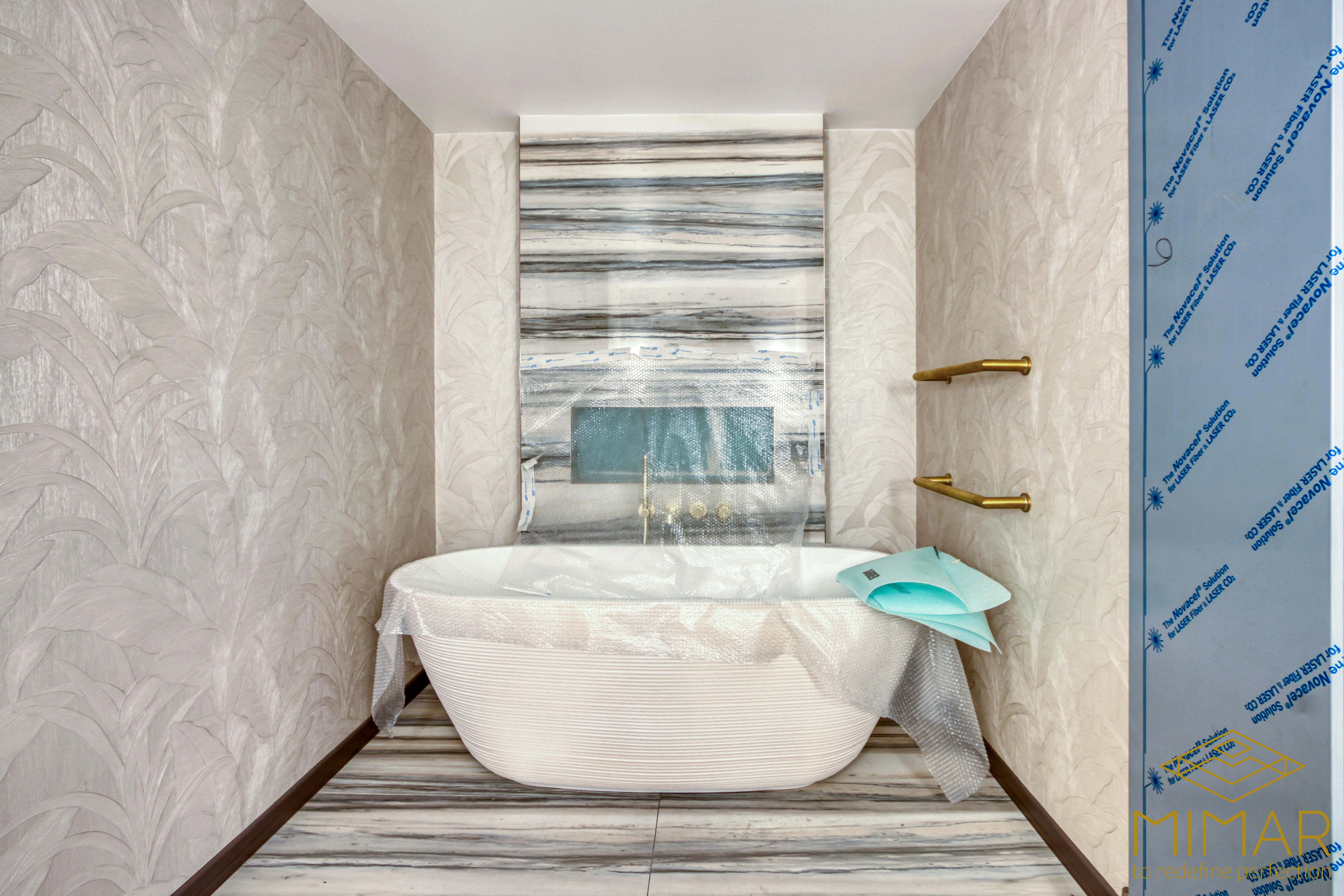 A modern bathroom under renovation featuring a freestanding bathtub with protective covering, marble tiles, subtle wallpaper, and gold fixtures.