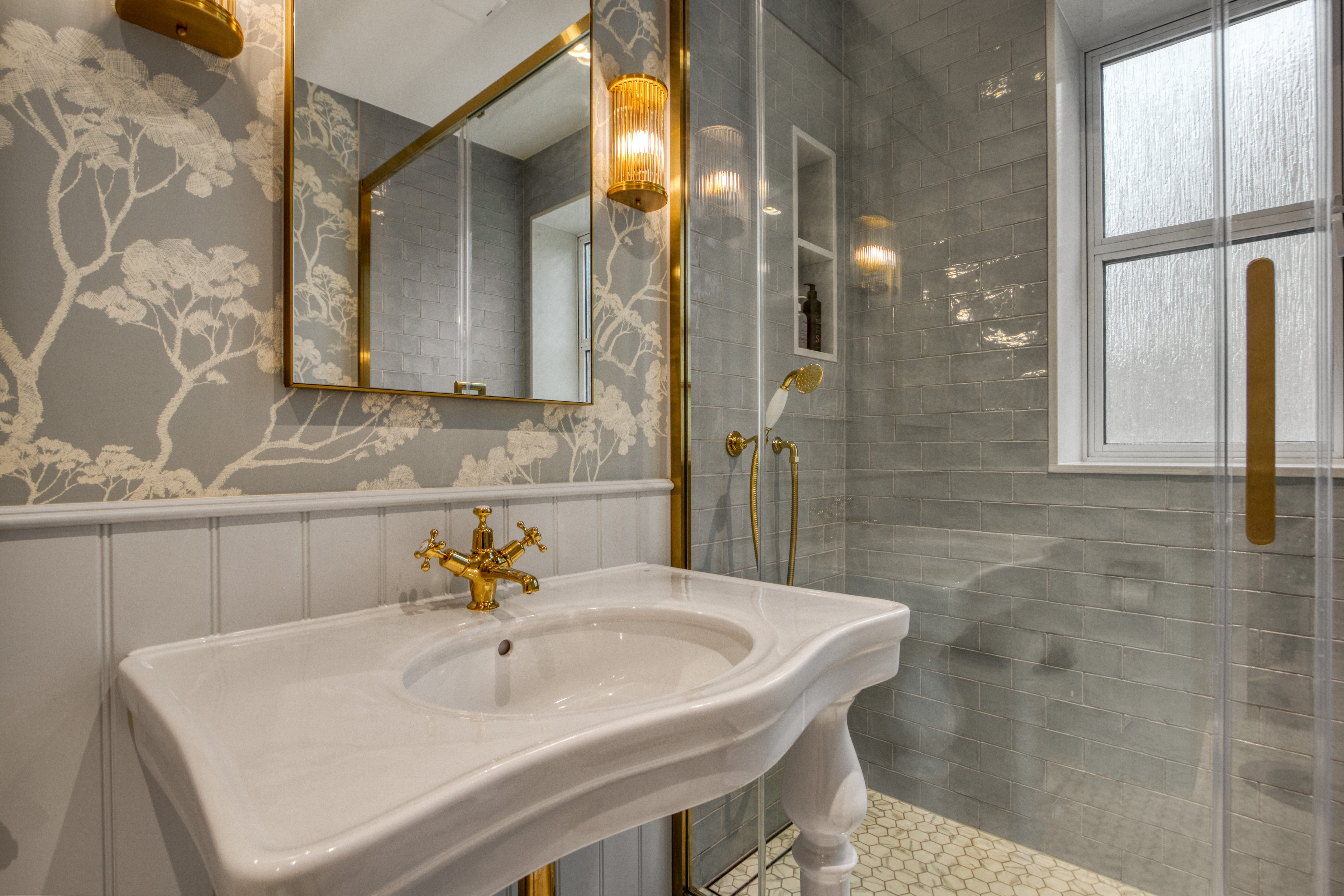 A modern bathroom with a vintage-style white ceramic sink, gold fixtures, patterned wallpaper, and a glass-enclosed shower area.