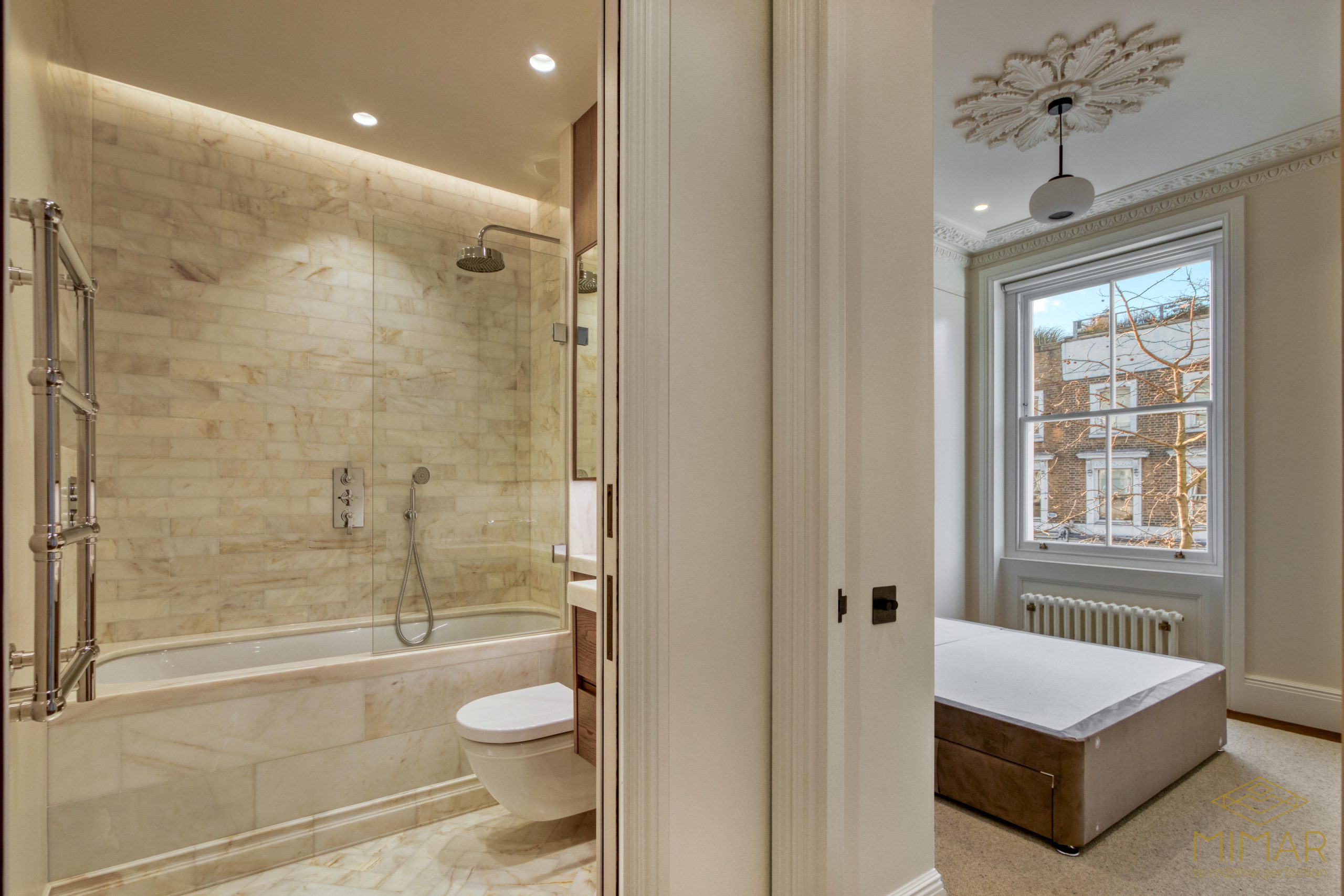A modern bathroom featuring a marble-tiled shower and bathtub area with glass enclosure, adjacent to a bright bedroom with elegant molding and large windows.