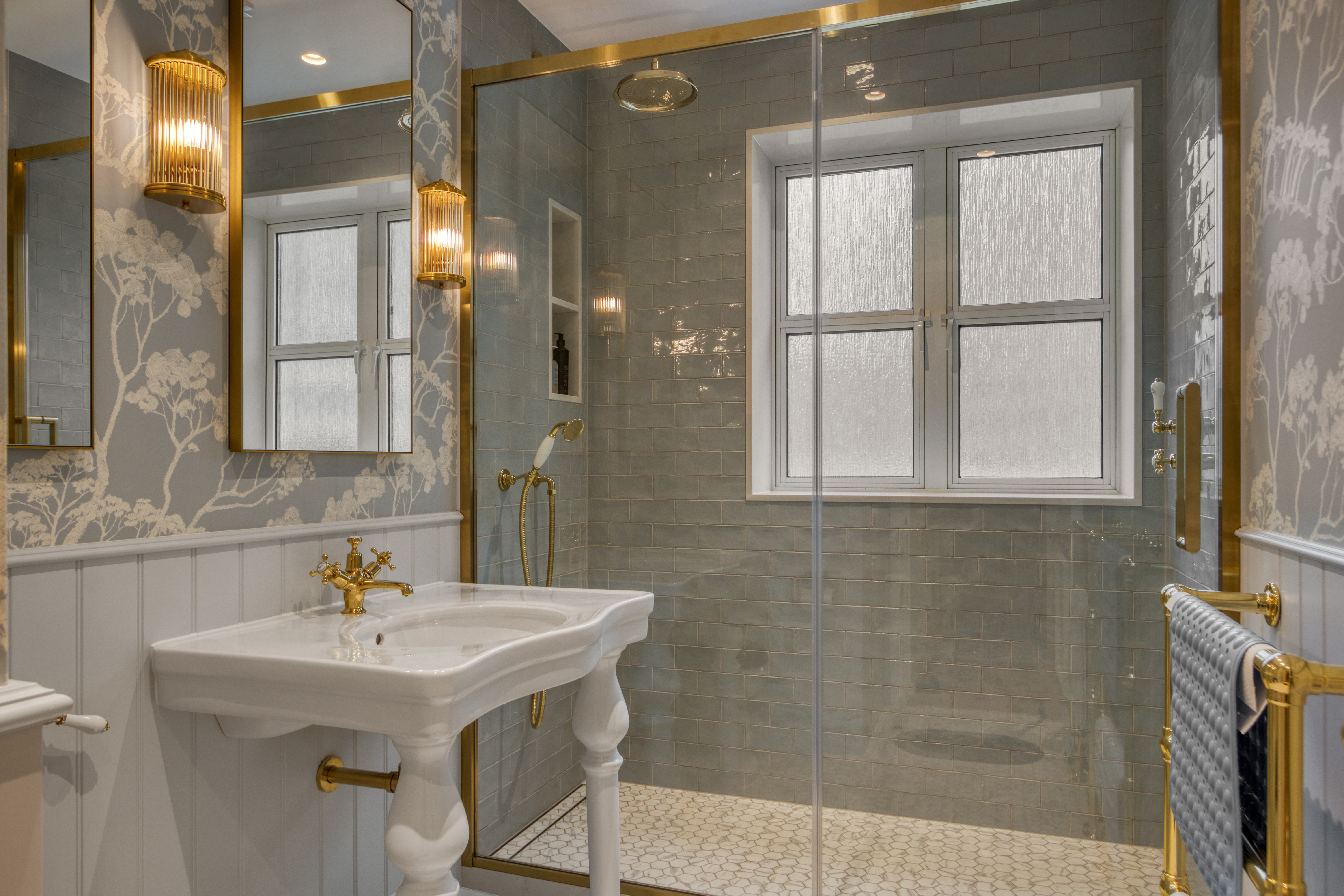 Elegant bathroom with a vintage-style sink, glass shower enclosure, and gold fixtures set against a floral wallpaper and tiled wall.