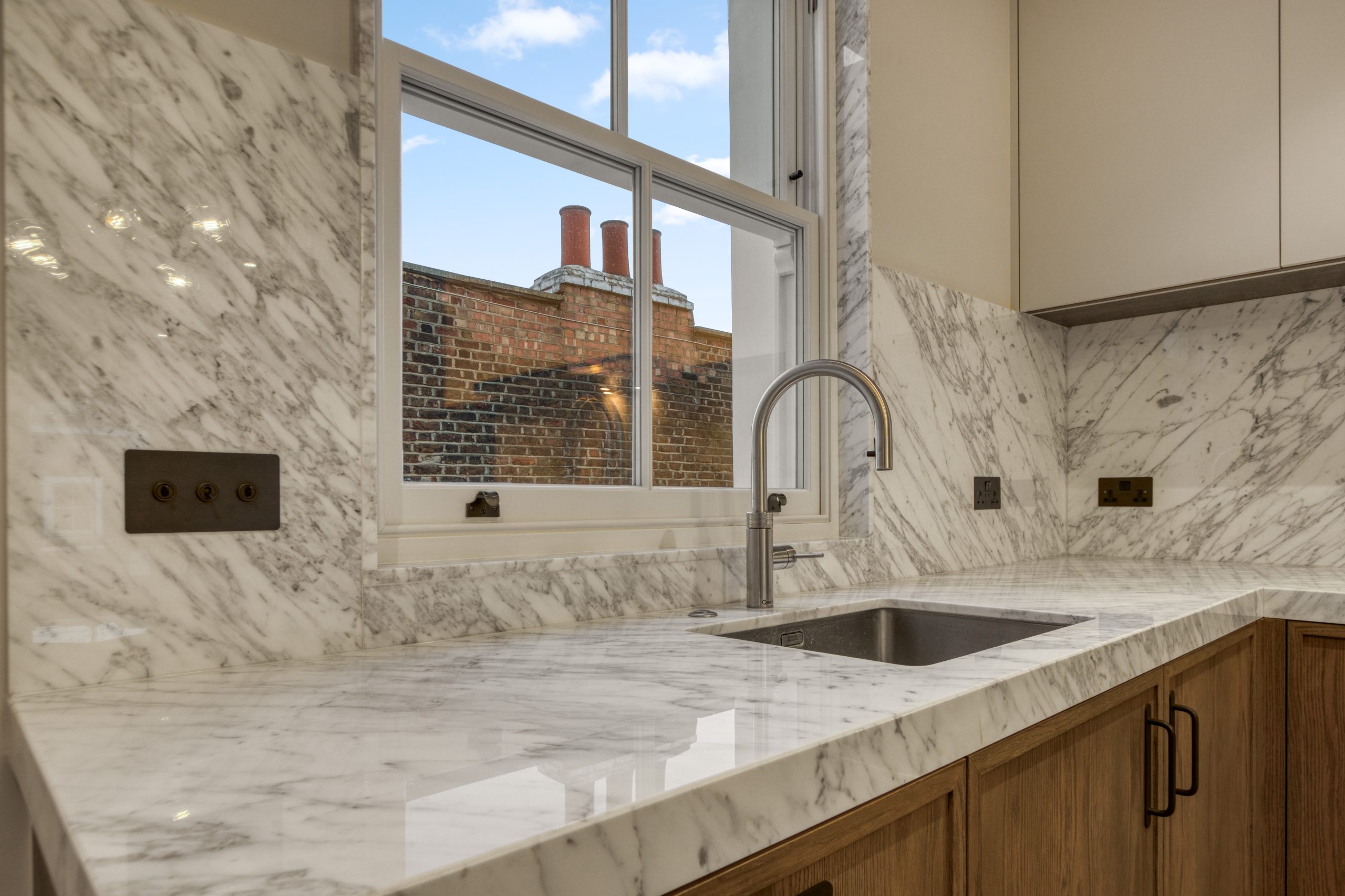 Modern kitchen featuring marble countertops, a stainless steel sink, and a window with a view of brick architecture.