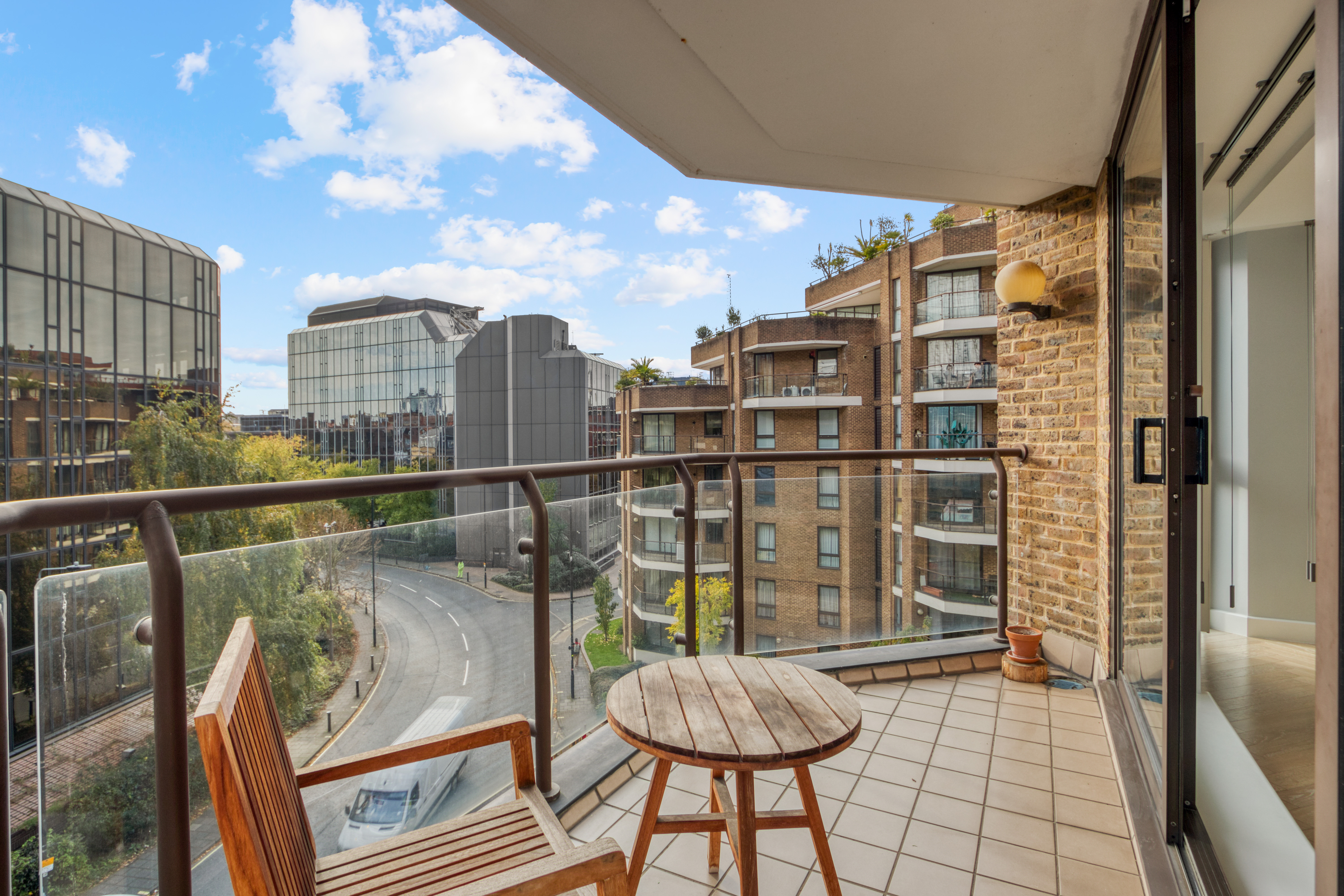 View from a modern balcony overlooking an urban area with surrounding buildings, a wooden chair, and small table.