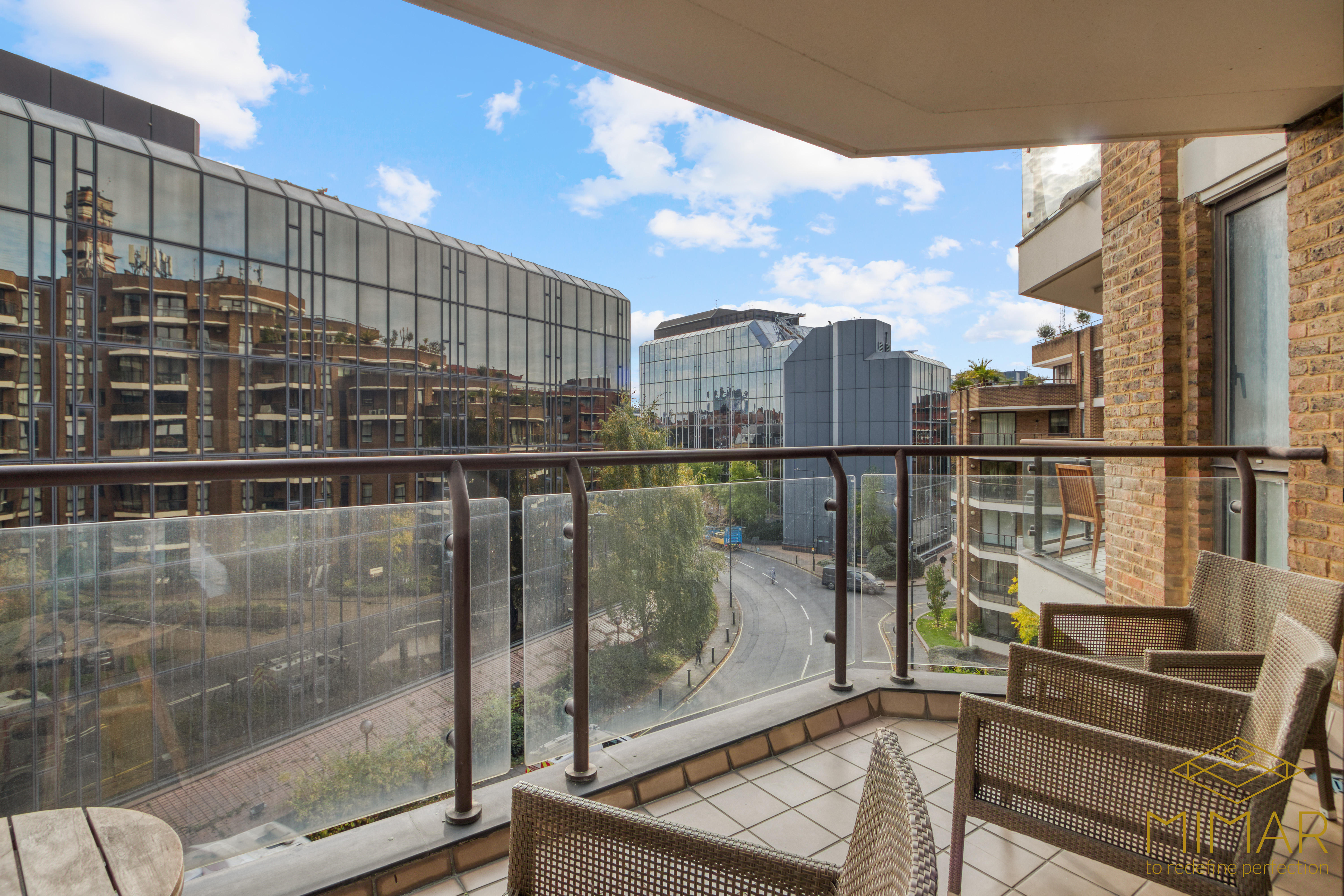 The image shows a modern balcony with glass railing and outdoor furniture, overlooking a street with modern buildings.