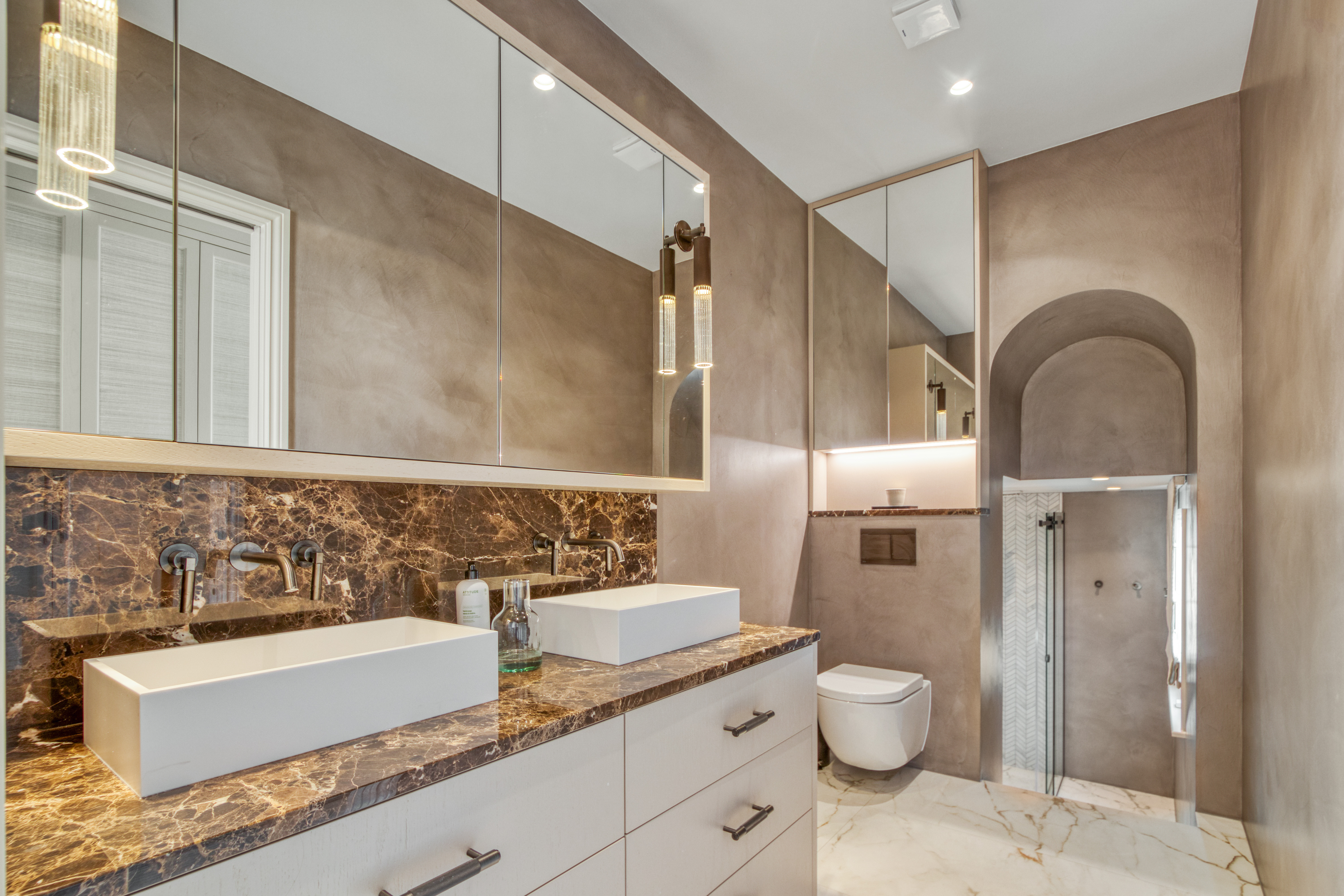 A modern bathroom featuring a double vanity with marble countertop, wall-mounted faucets, and a large mirror above. The space includes a toilet and a walk-in shower with elegant marble and neutral tones.