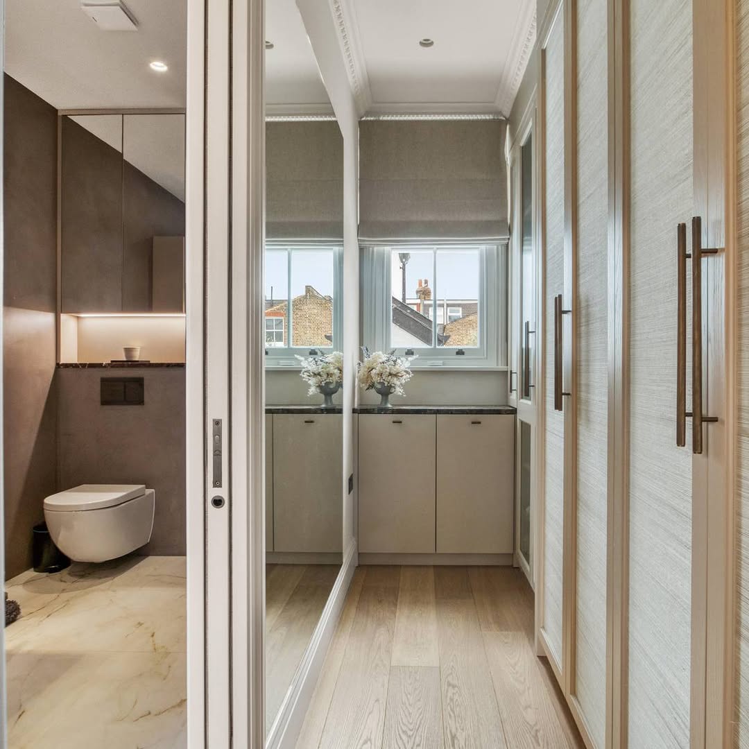 Modern bathroom featuring a wall-mounted toilet, large mirror, marble flooring, and integrated cabinetry next to a window with a view of outside buildings.