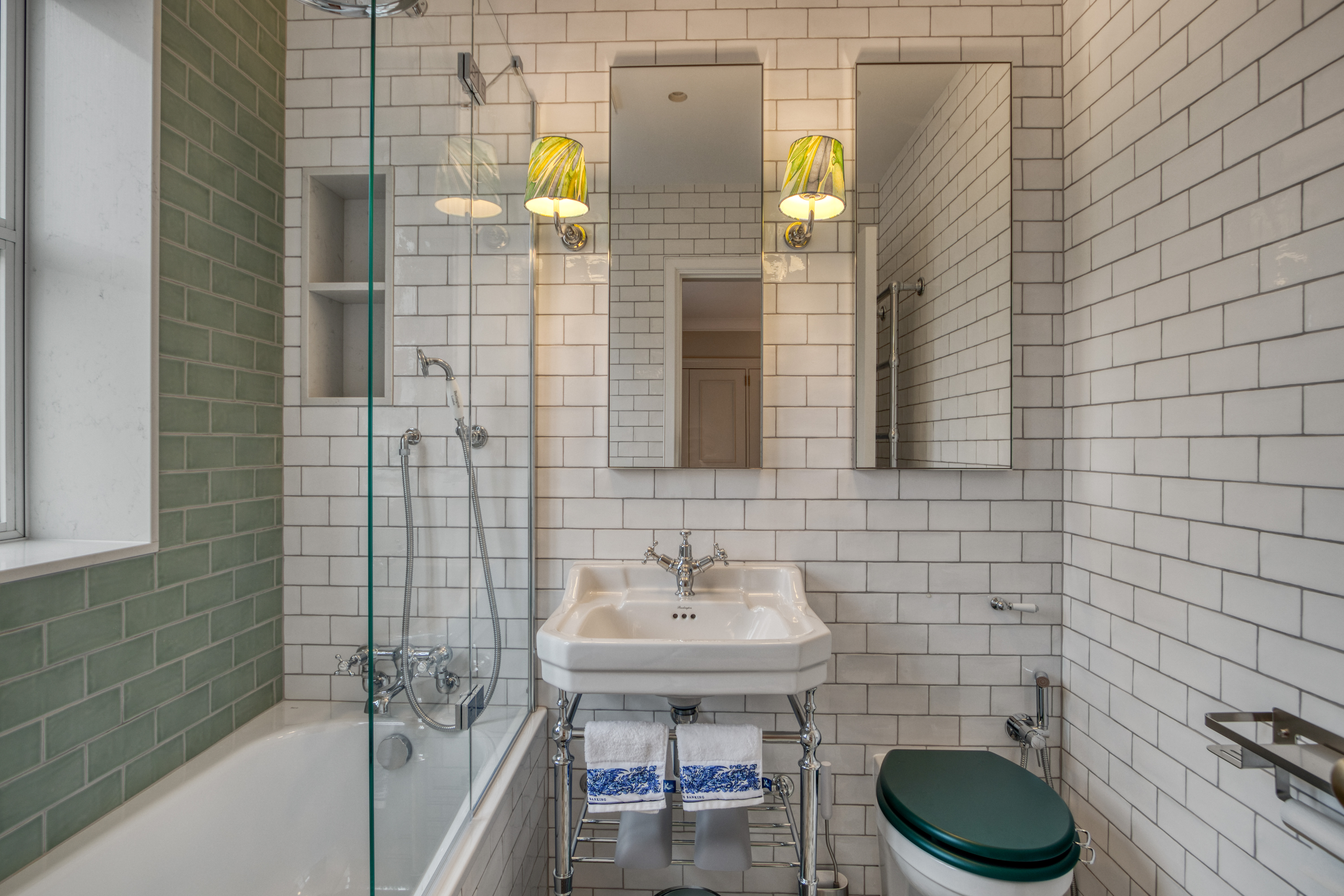 A modern bathroom featuring white subway tiles, a glass-enclosed shower-bathtub combination, a pedestal sink, and a wall-mounted toilet with traditional fixtures.