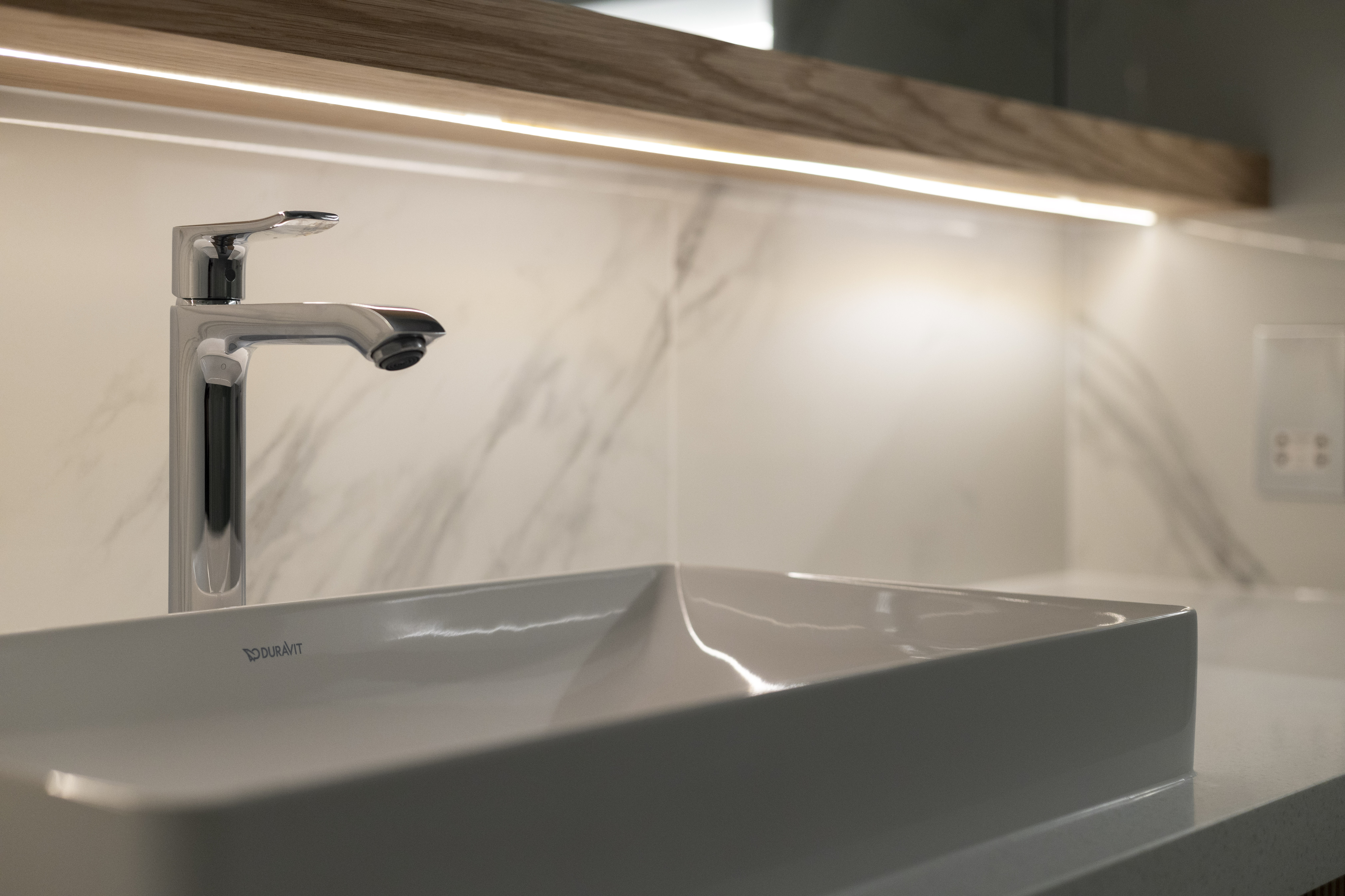 A modern bathroom sink with a sleek chrome faucet and marble backsplash, illuminated by under-cabinet lighting.