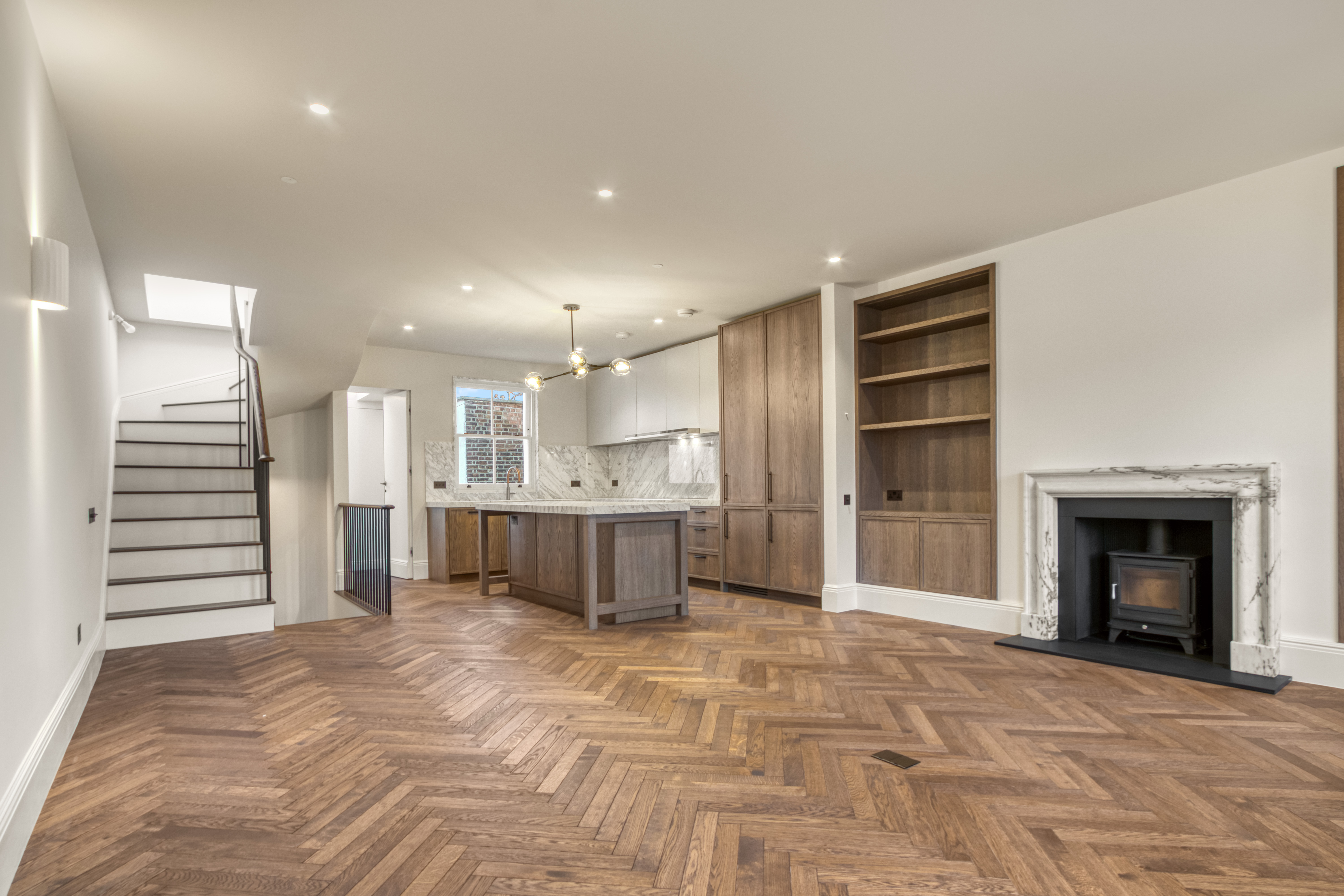 Open-plan kitchen and living area with herringbone wooden flooring, a modern kitchen island, fireplace, built-in shelves, and staircase.