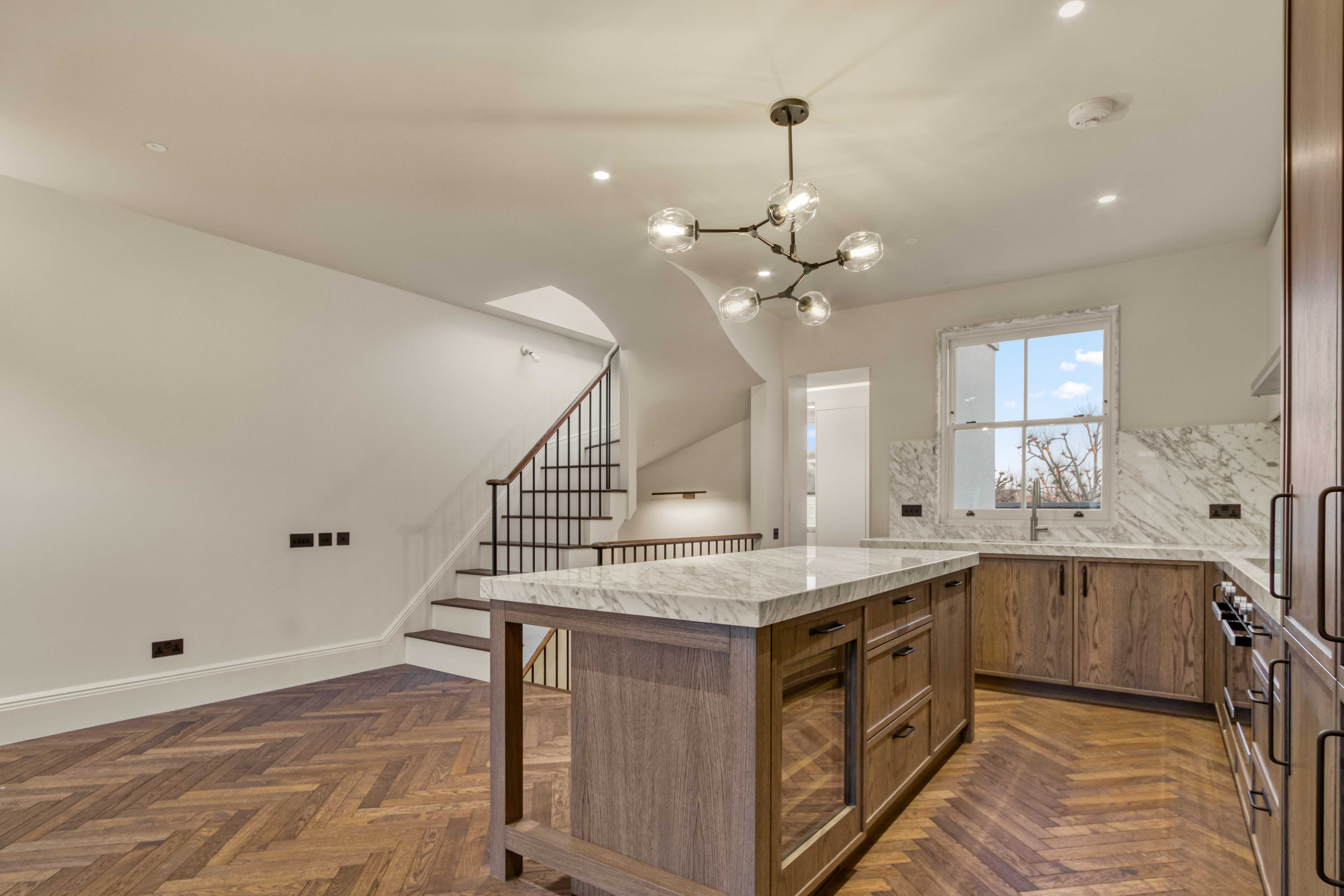 A modern kitchen with wooden cabinetry, marble countertops, and island, featuring a contemporary light fixture and herringbone hardwood flooring.