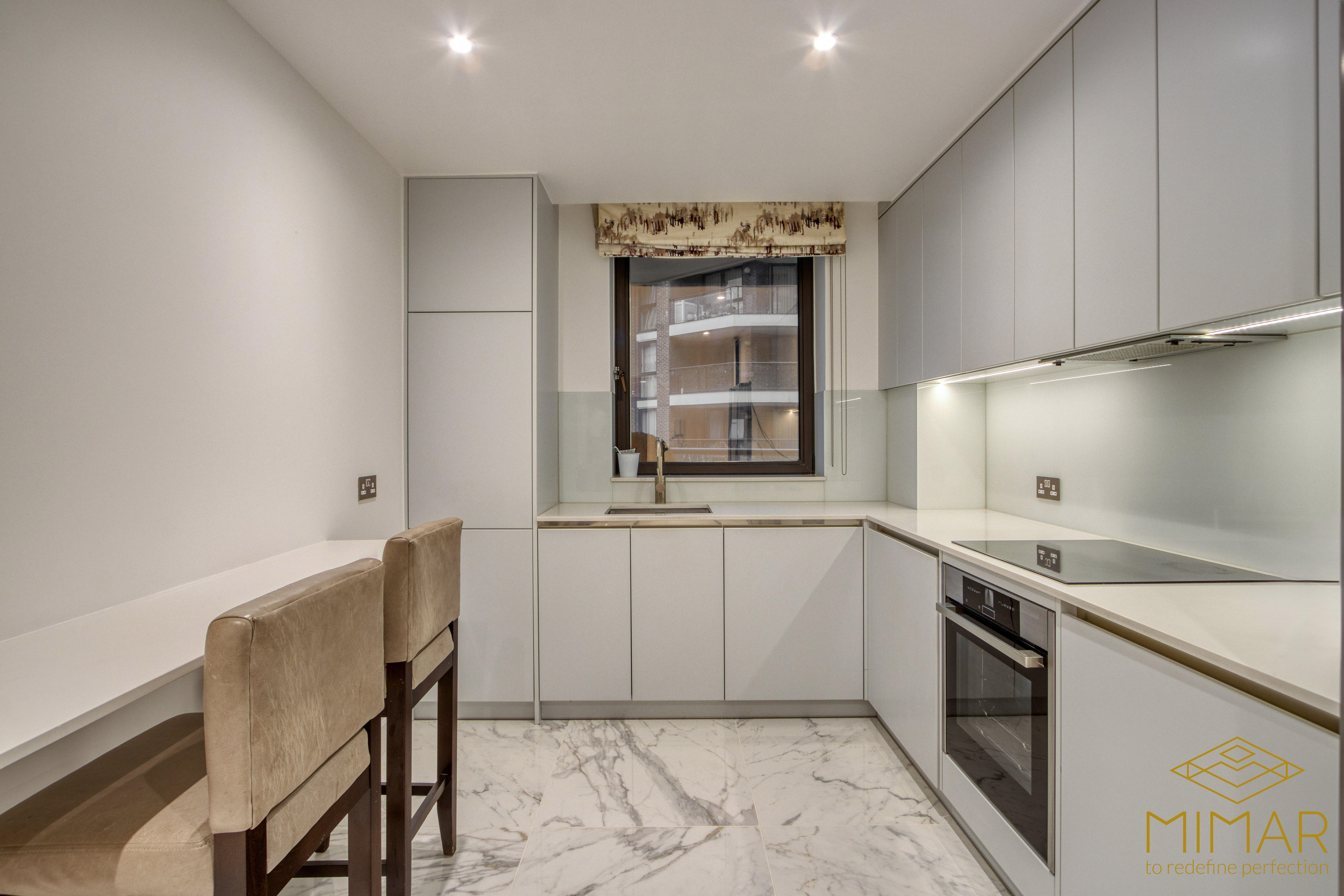 A modern kitchen featuring white cabinets, a built-in oven, marble flooring, and a sleek countertop with two bar stools.