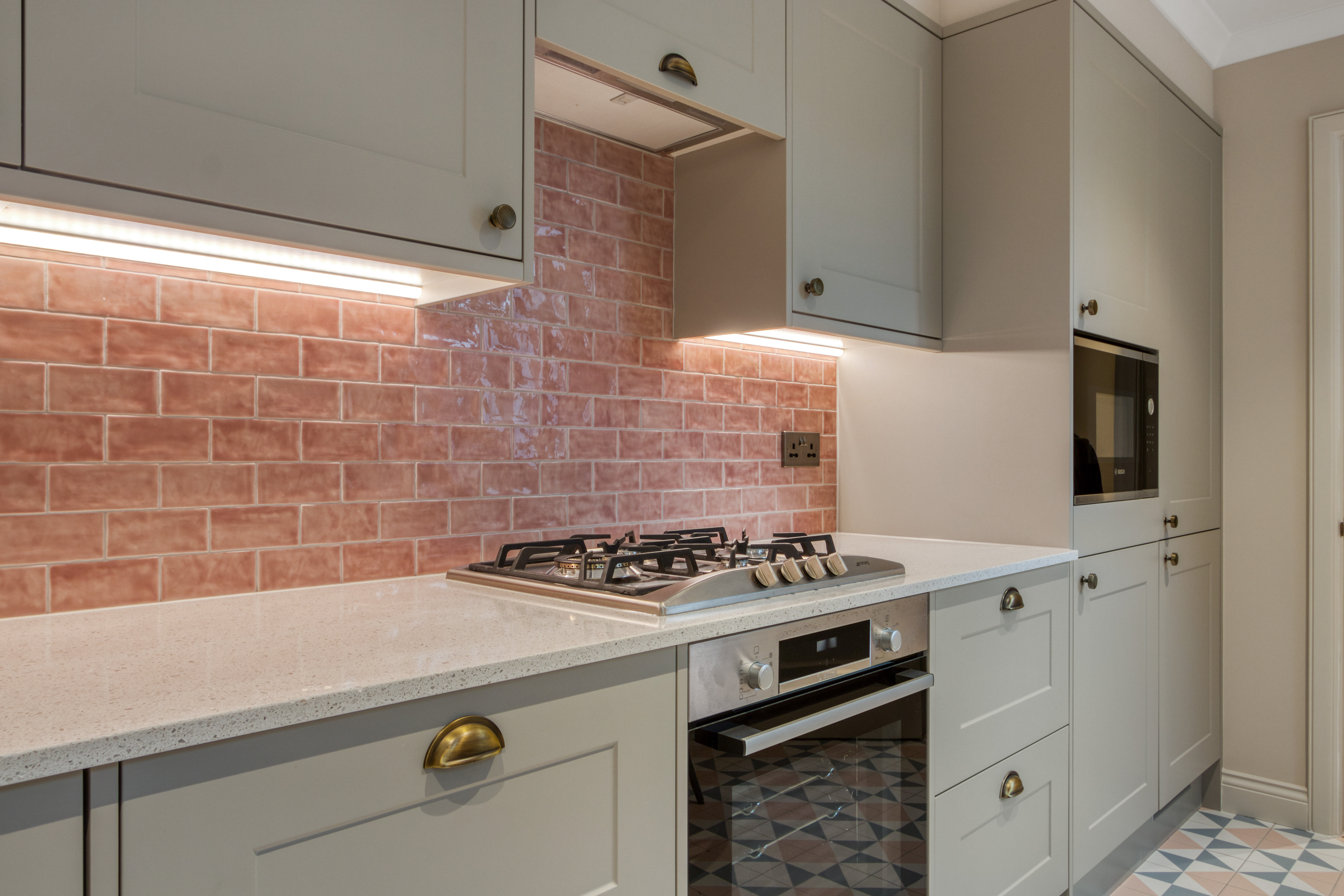 A modern kitchen with light-colored cabinets, a built-in oven, and a gas stove, featuring a pink tile backsplash and under-cabinet lighting.