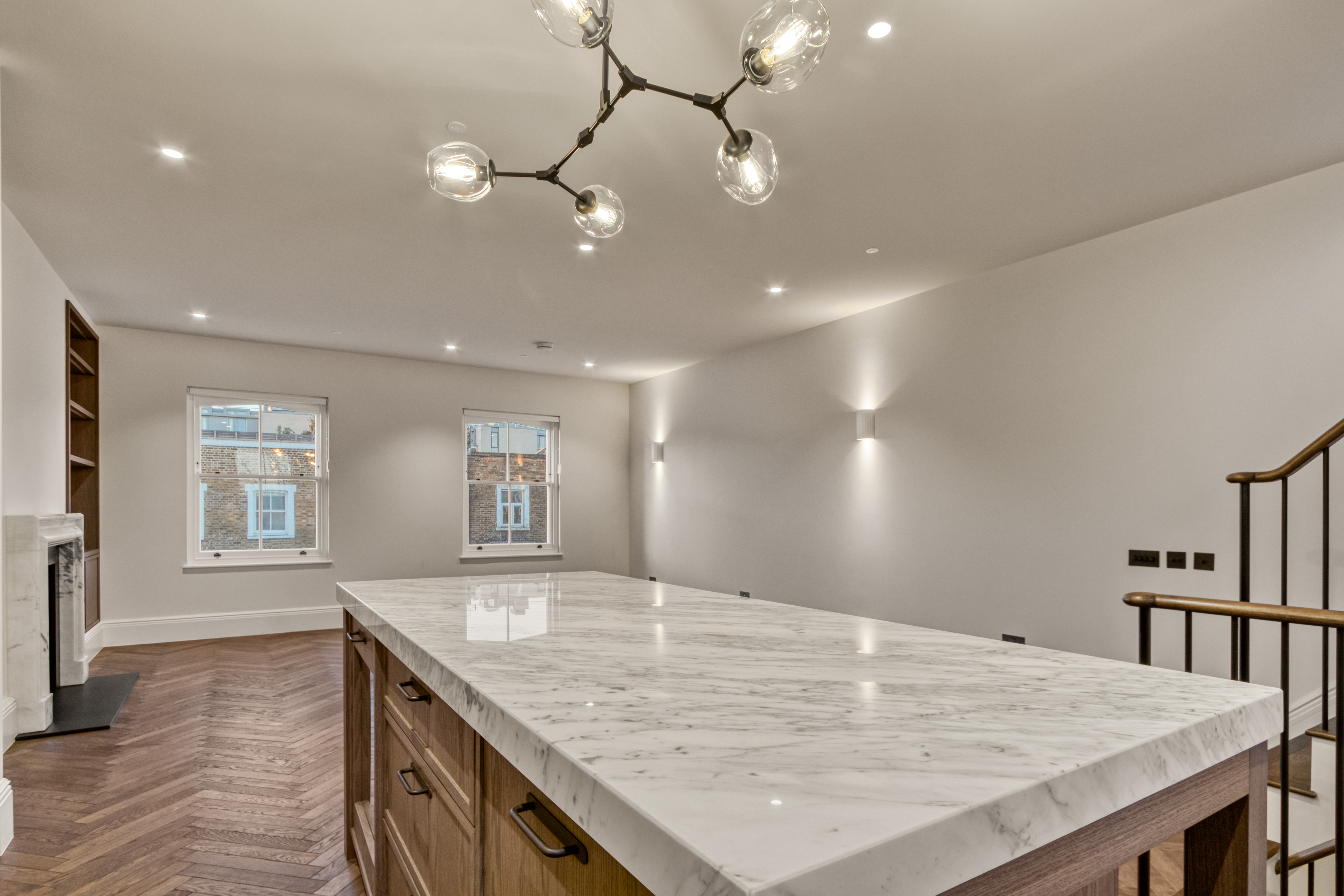 A modern kitchen interior with a large marble island, wooden cabinets, and contemporary lighting fixtures. The room features wooden flooring and a staircase with a metal railing.