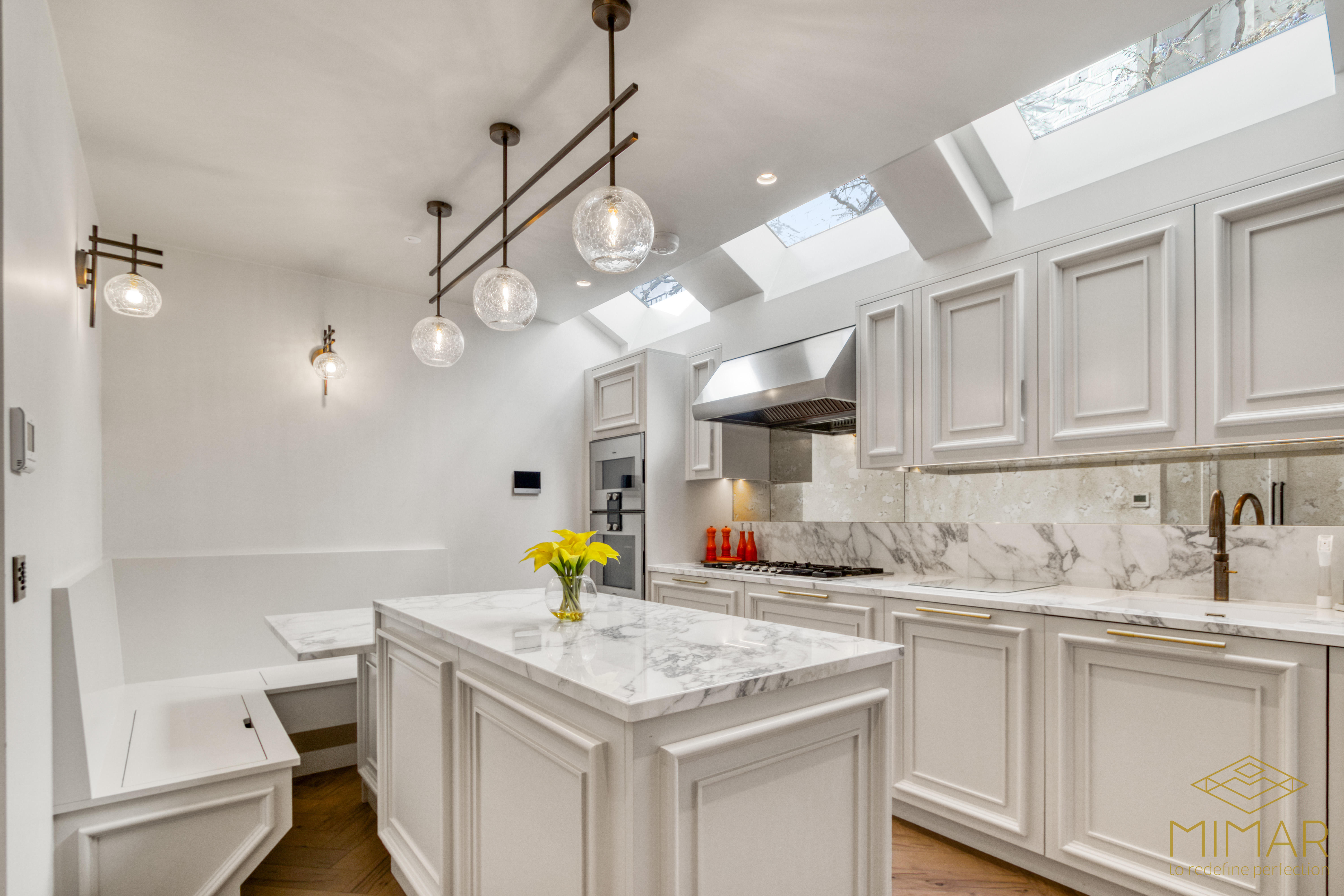 A modern kitchen featuring white cabinetry, marble countertops, and stainless steel appliances under skylights with pendant lighting.