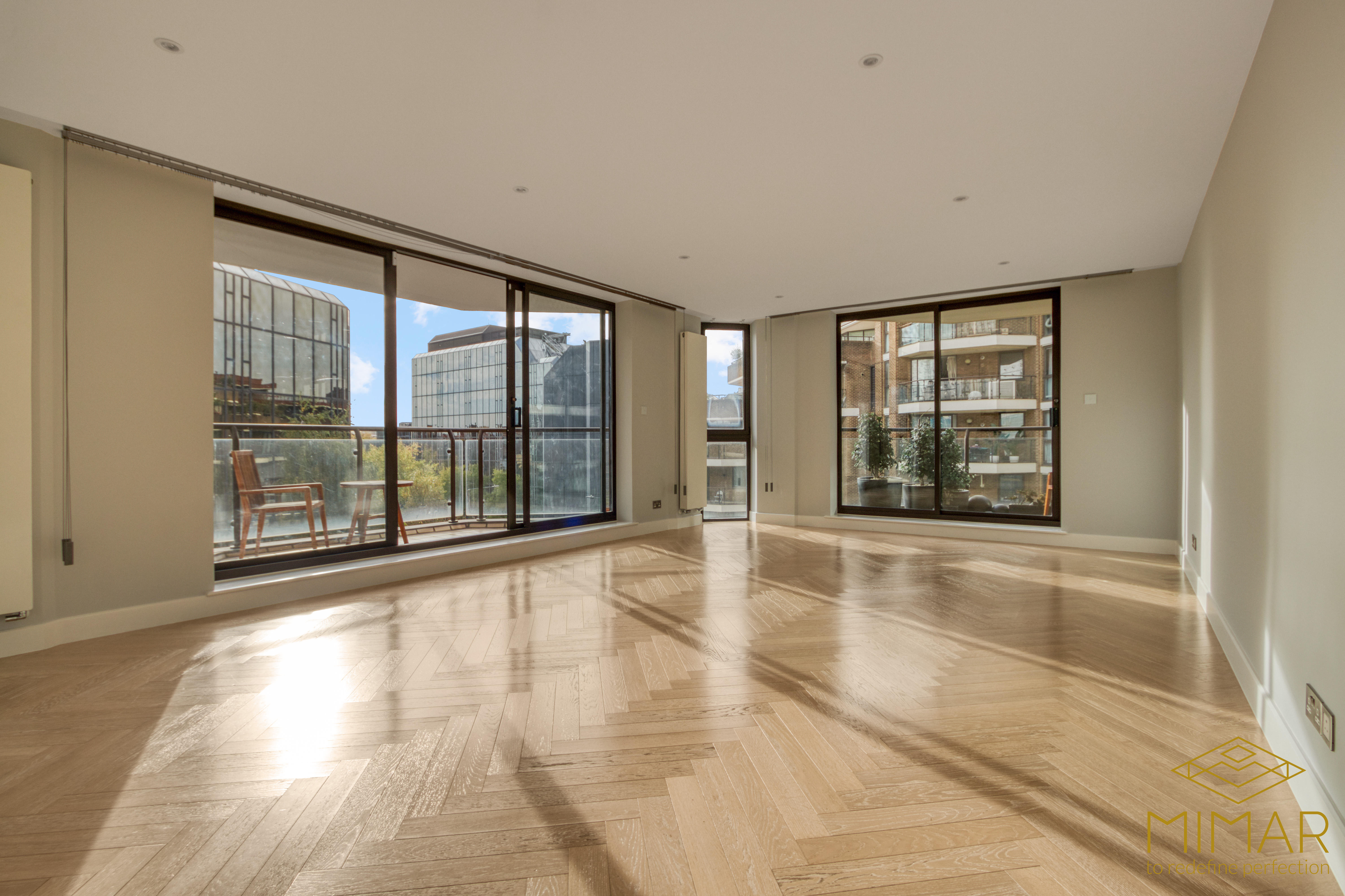 Spacious, modern apartment interior with large windows and wooden flooring, offering views of surrounding city buildings.