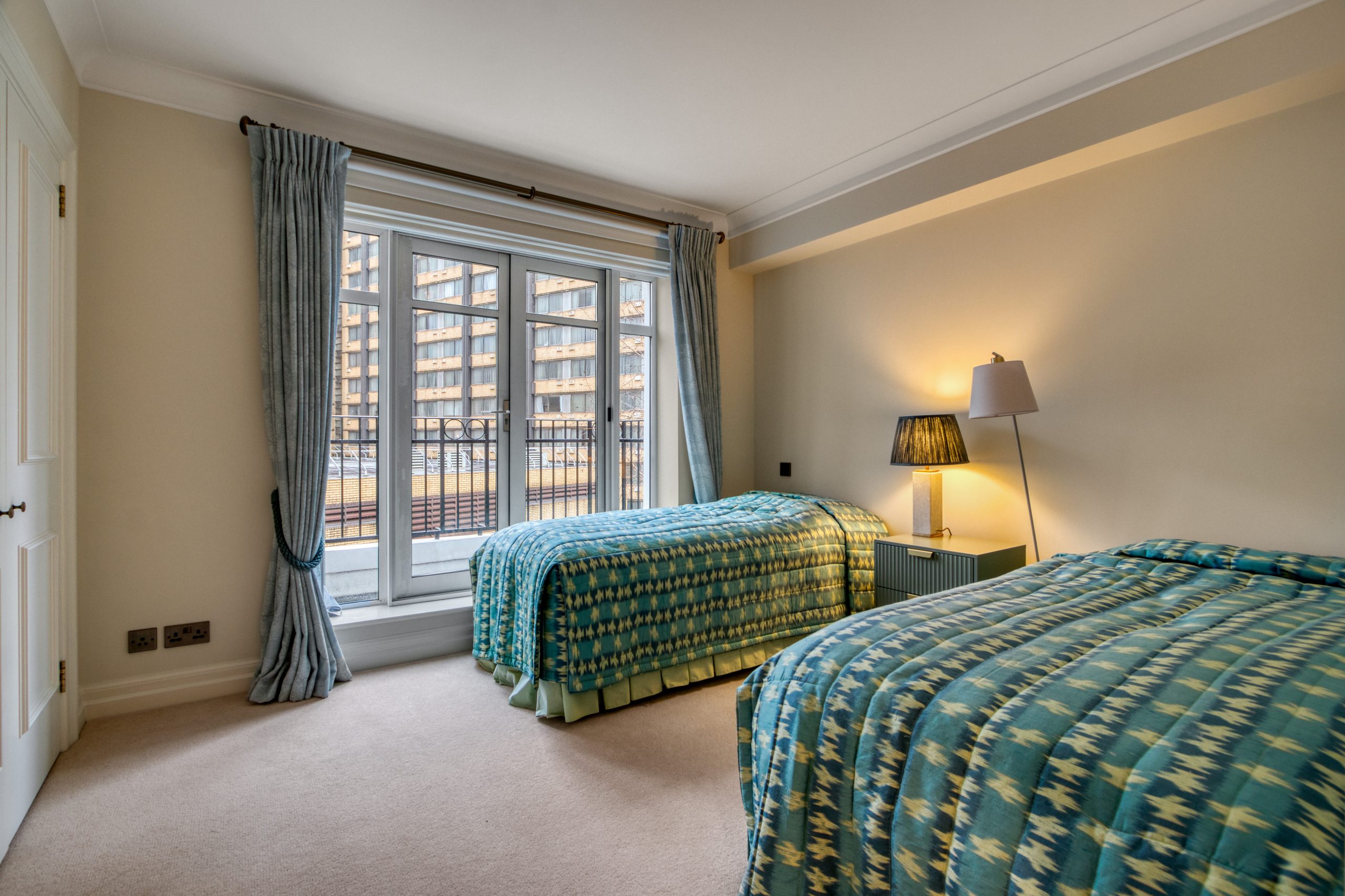A newly refurbished bedroom featuring two single beds, a window with curtains, and a lamp on a bedside table.