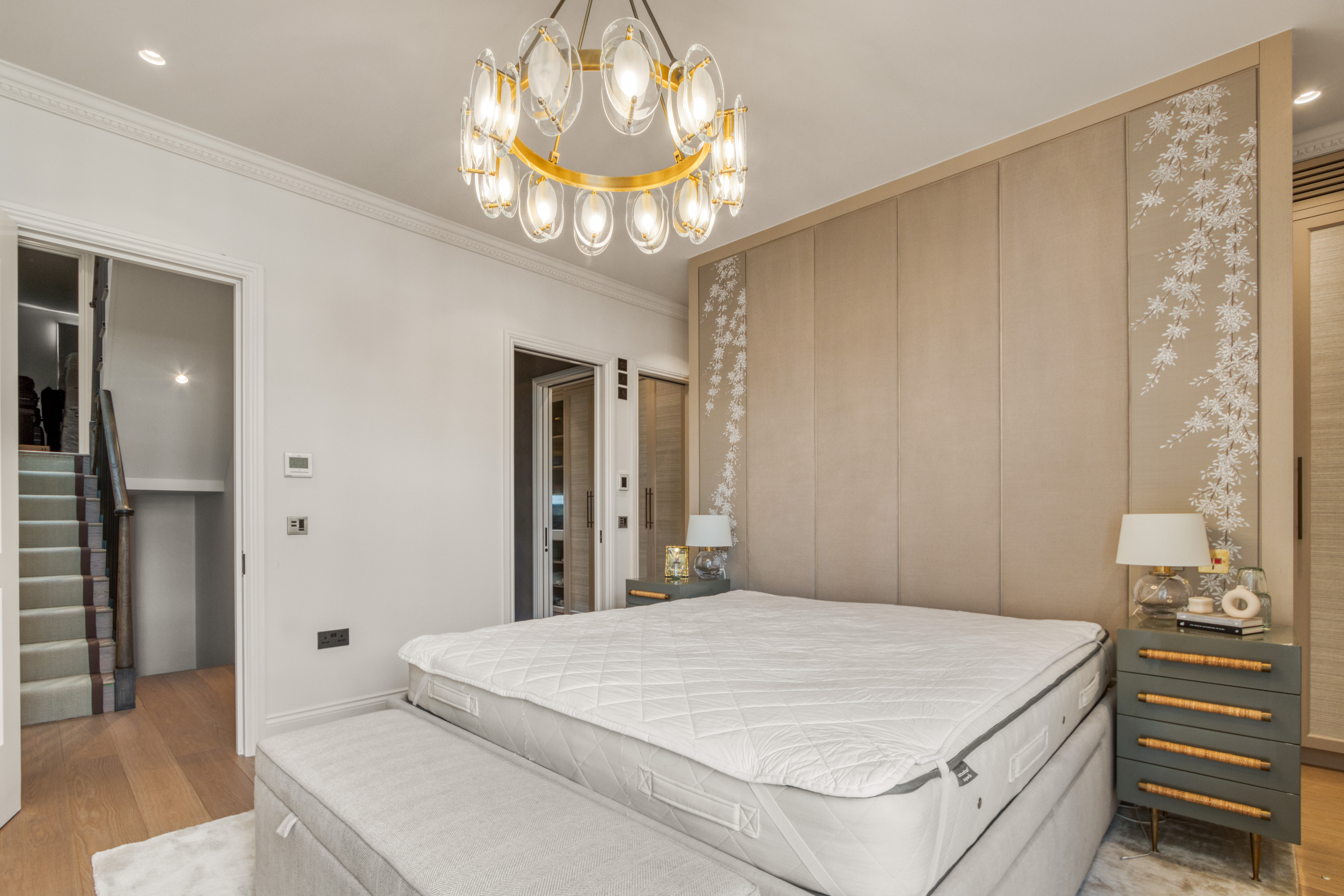 A modern bedroom with a large bed, decorative padded wall panels, stylish chandelier, and side tables, showcasing a contemporary refurbishment.