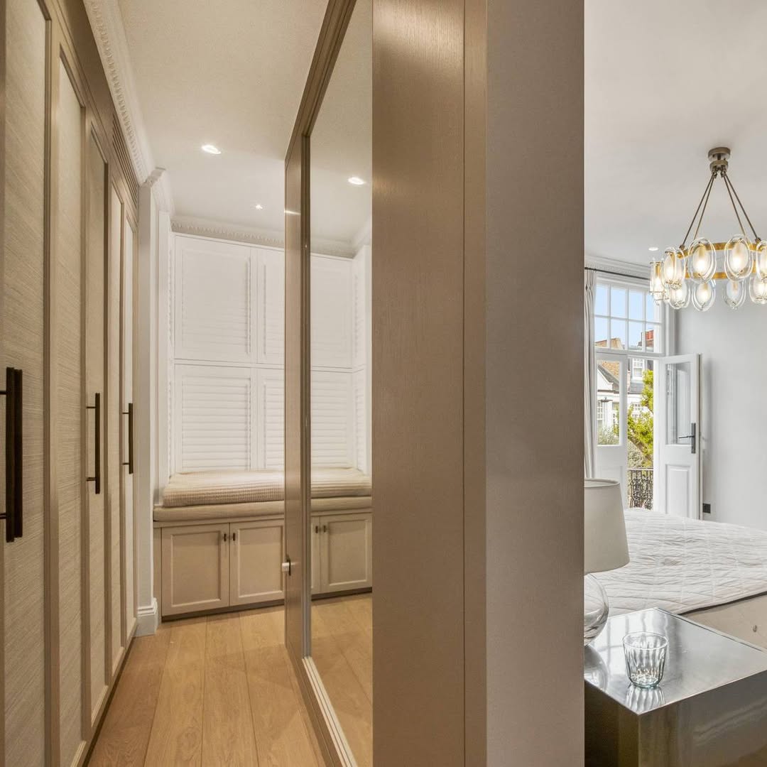 Interior view of a modern bedroom with built-in wardrobes and large windows, featuring a chandelier and a reflection in a mirrored door.