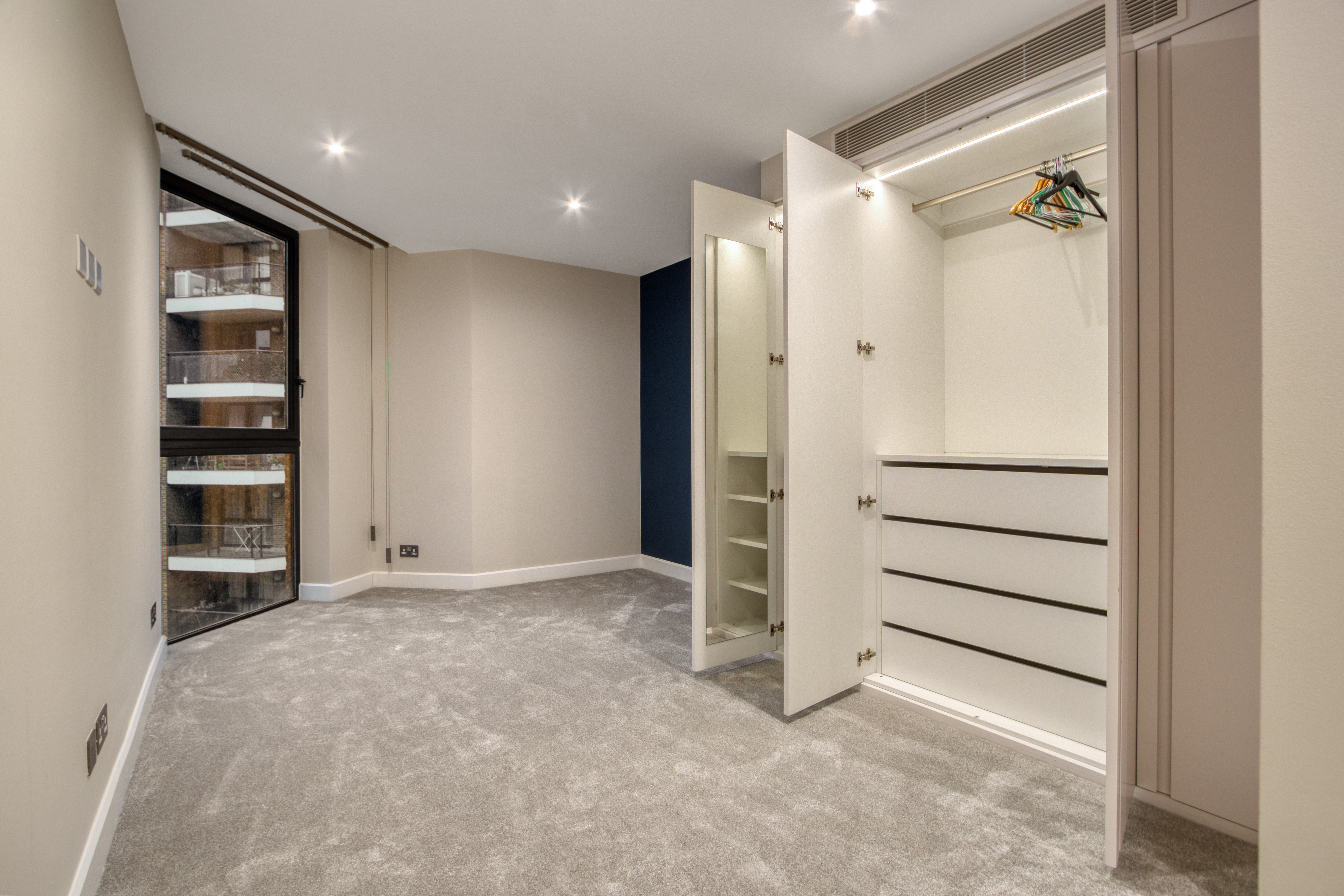 A newly refurbished bedroom with a built-in wardrobe featuring open doors, drawers, and hanging rail, and large windows offering a view of a balcony.