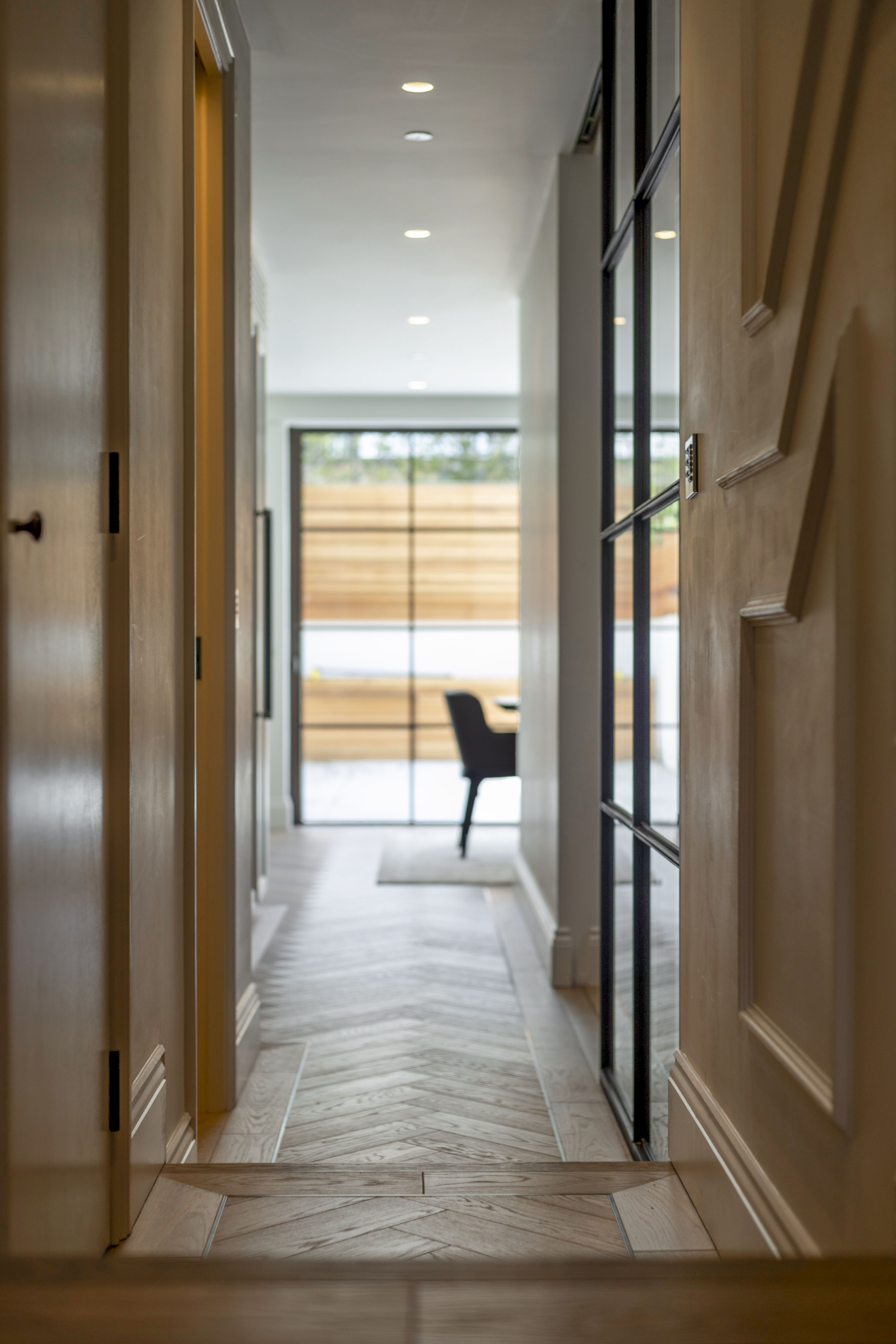 A modern and sleek hallway with wooden flooring, recessed lighting, and a view leading to a dining area with large glass doors.