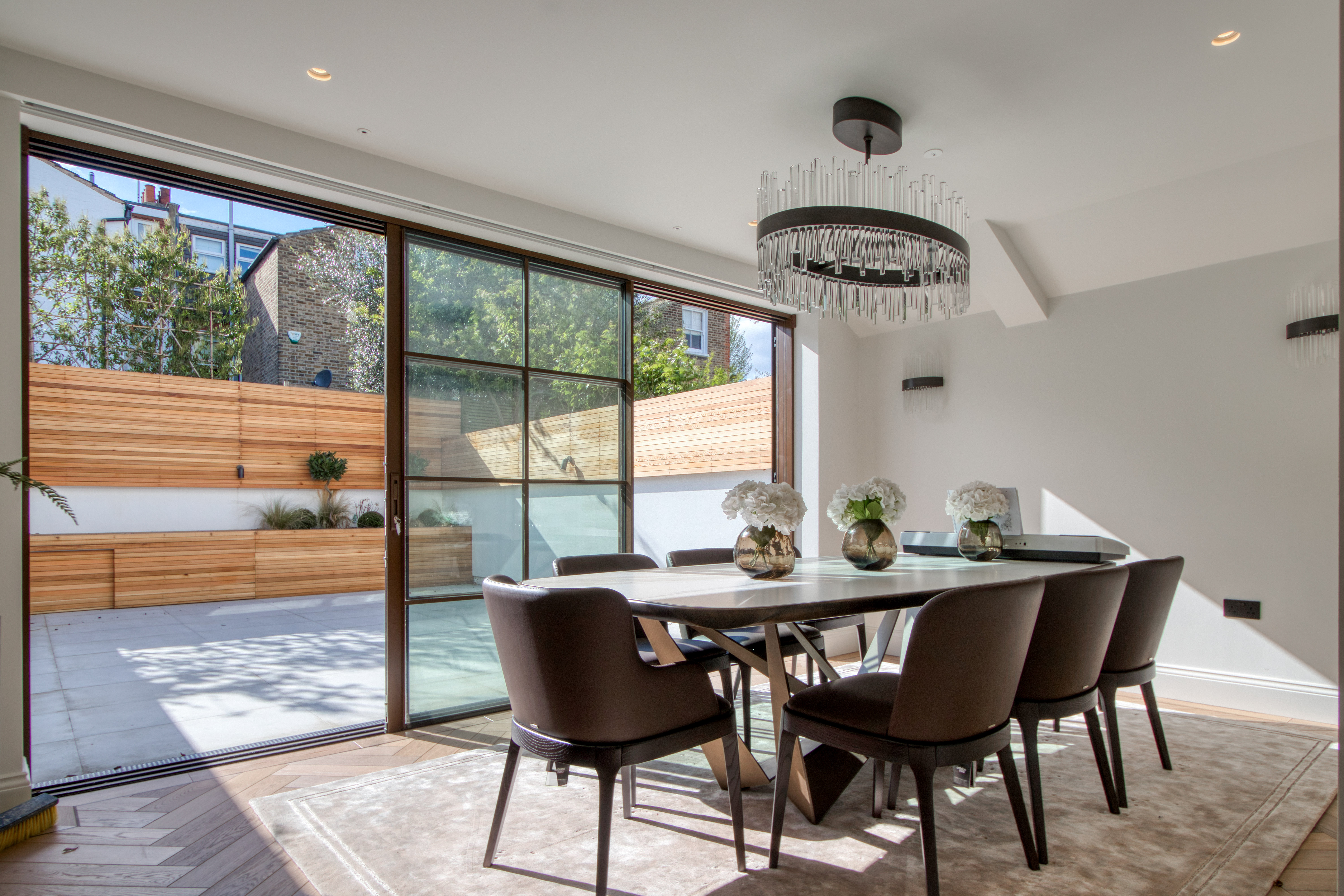 Modern dining room with large glass doors opening to a patio, featuring a contemporary chandelier and elegant decor.