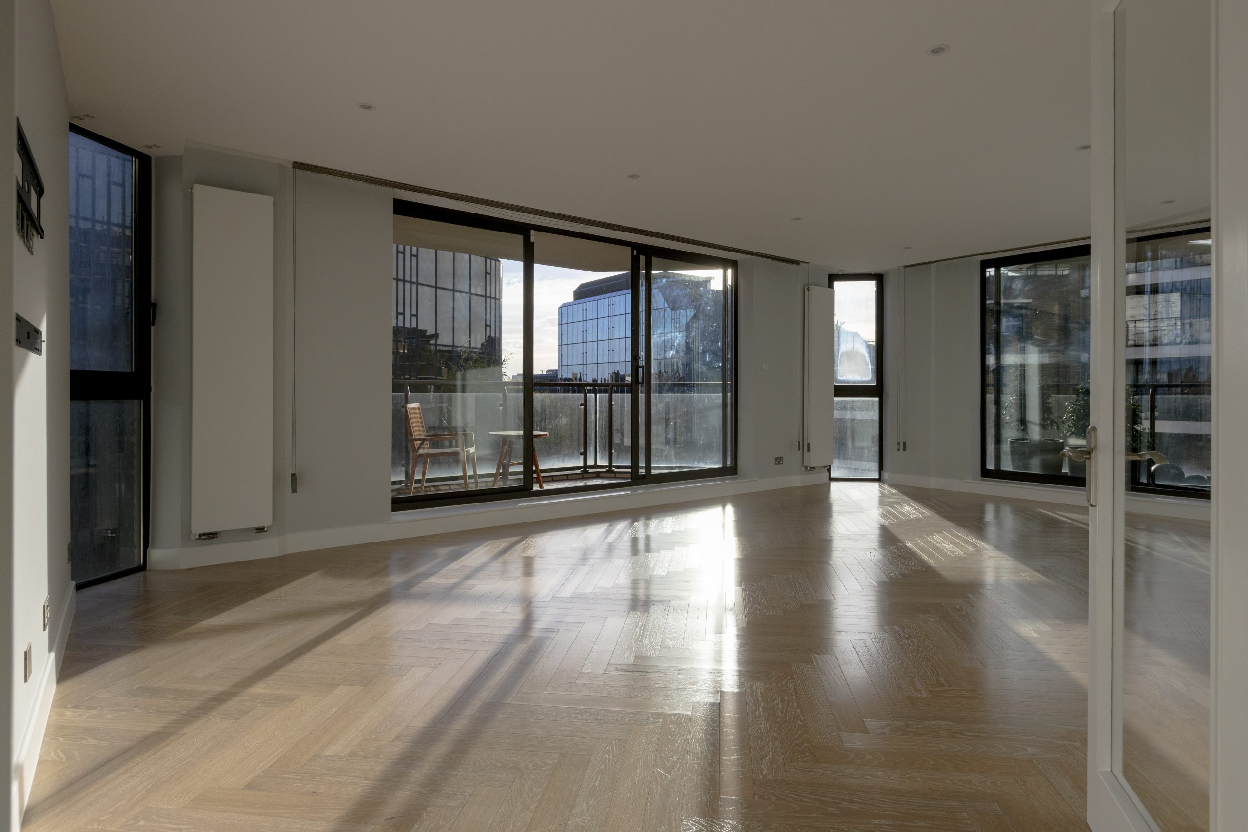 Spacious, modern living room with wooden herringbone flooring and large glass doors leading to a balcony.