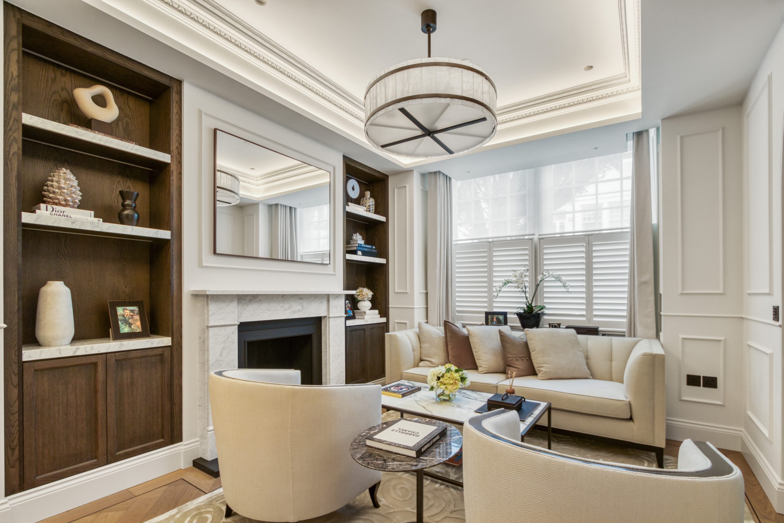 A well-designed living room featuring custom carpentry shelving, an elegant mirror above a marble fireplace, and contemporary furnishings with a beige color palette.