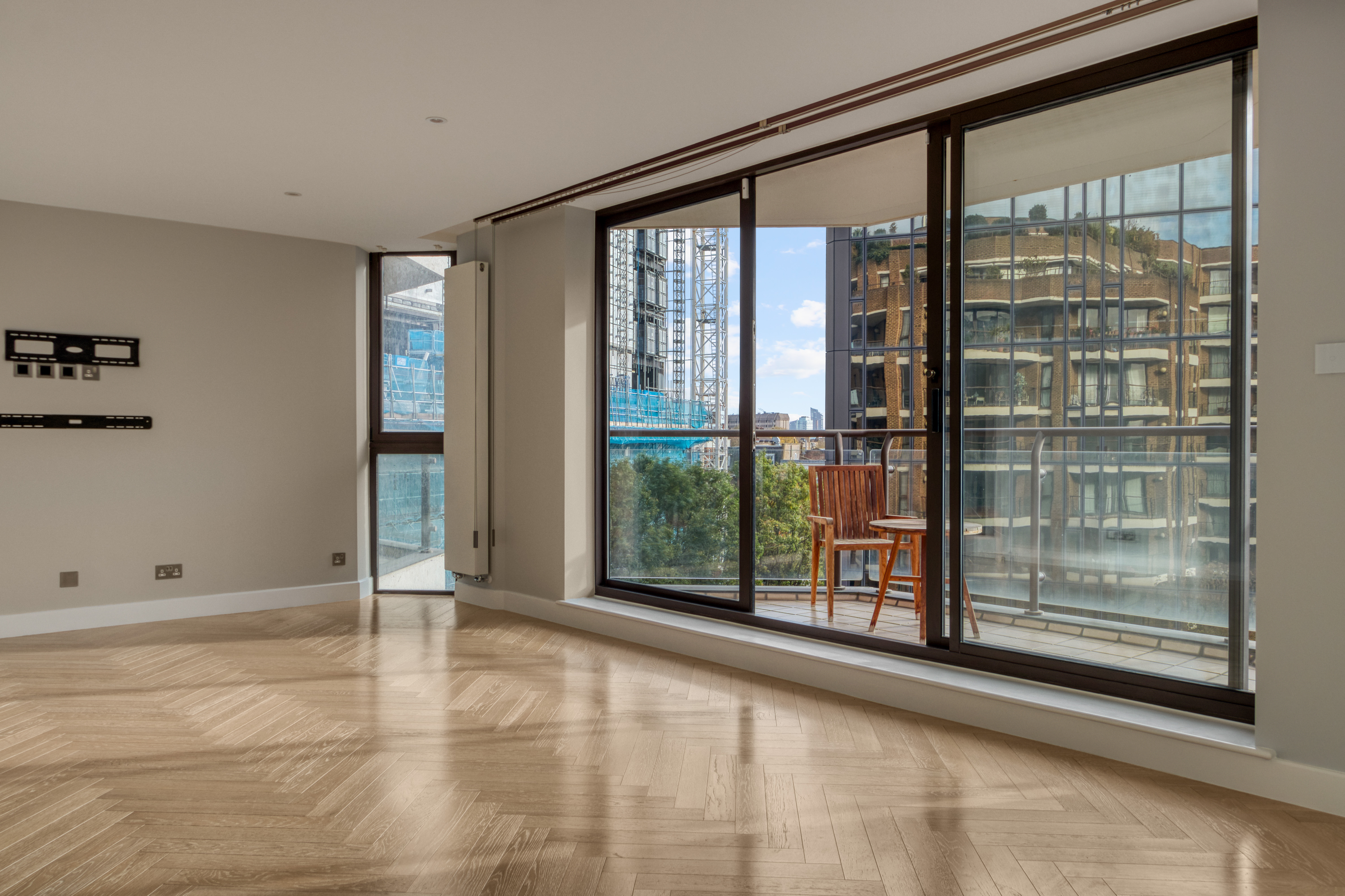 A modern living room with wooden flooring and large sliding glass doors leading to a balcony, showcasing an urban view.