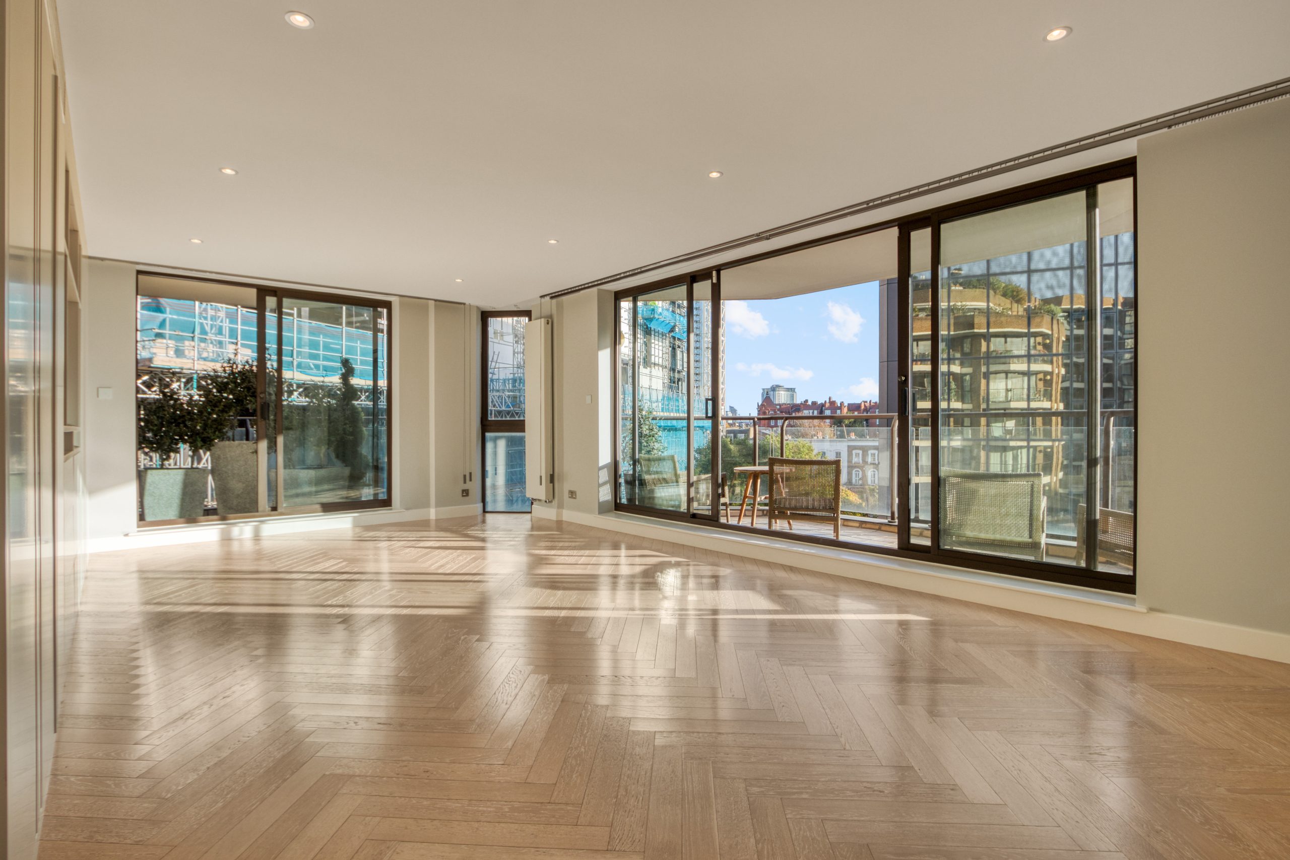 Spacious open-plan living area with large glass windows and wooden flooring, overlooking a balcony with city views.