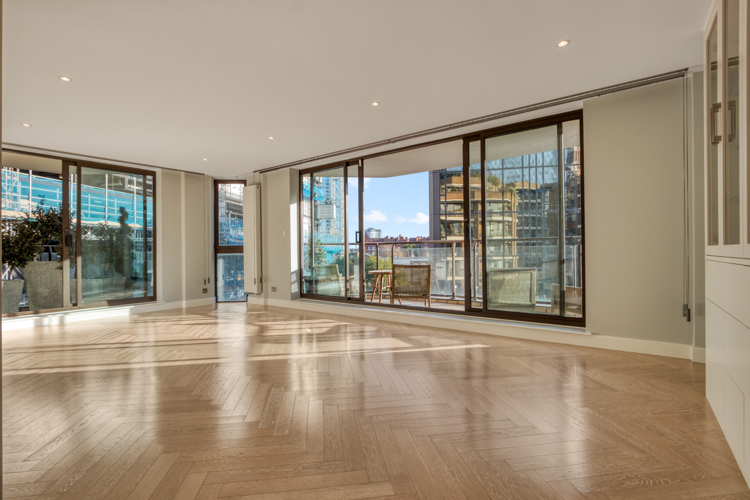 An open-plan living area with large floor-to-ceiling windows and modern flooring, offering a view of nearby buildings.