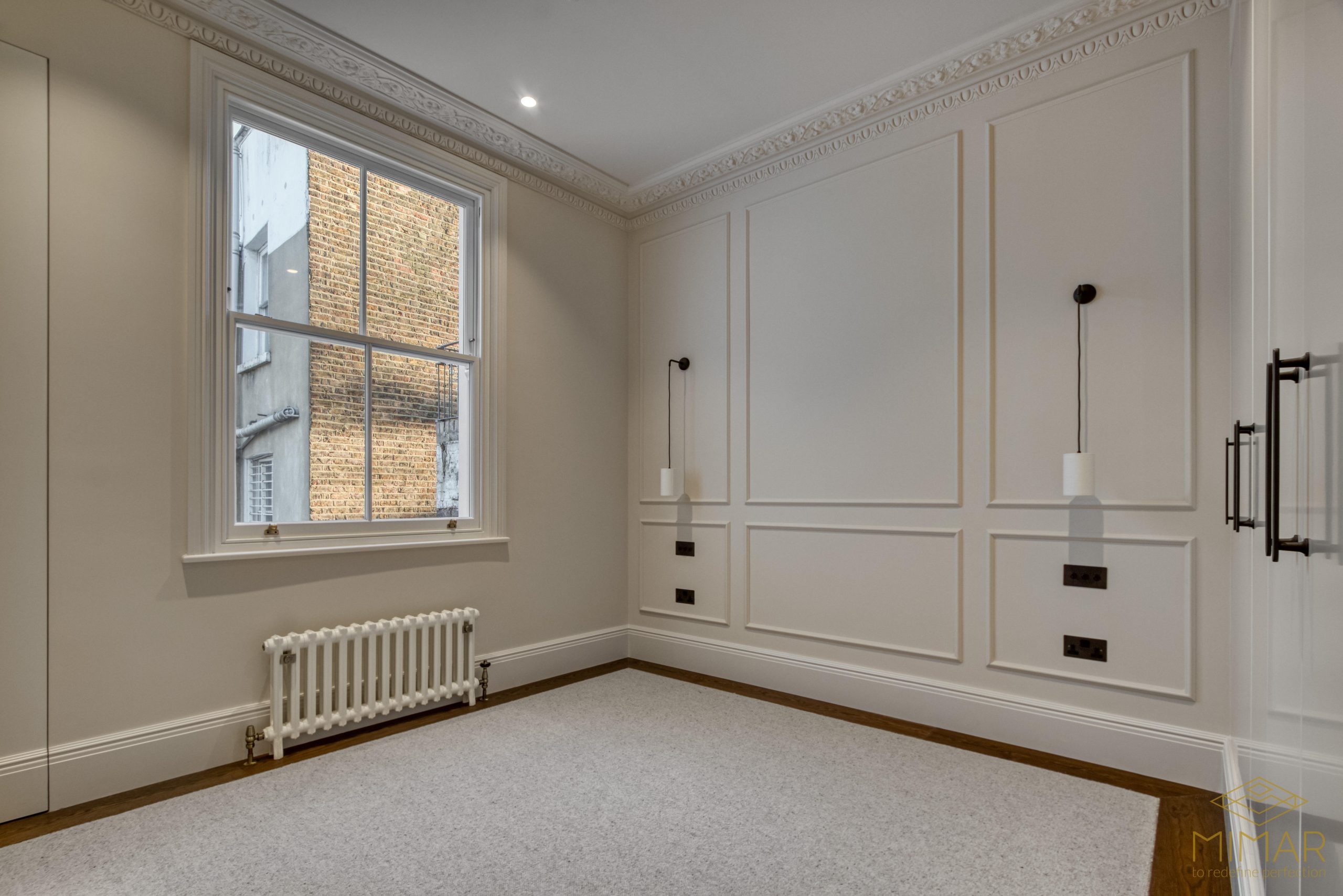A newly refurbished room featuring elegant wall paneling, a large window with a view of an exterior brick wall, and a traditional radiator beneath the window.